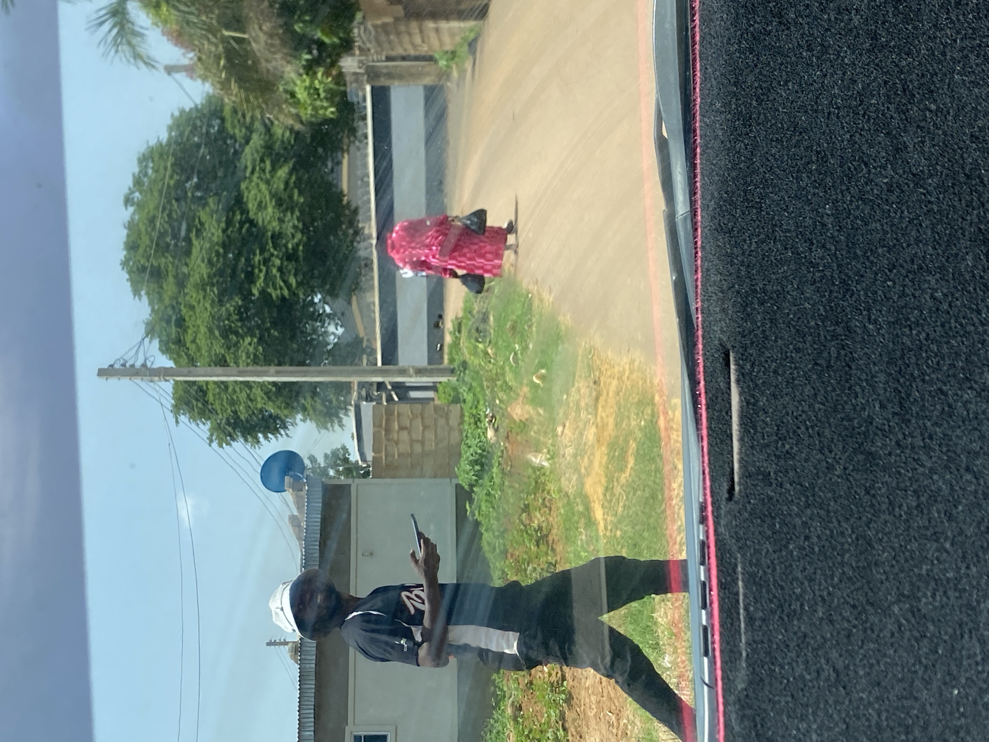 A man wearing a white bucket hat and a black t-shirt with white and black trim walks on a dirt road in Bar Jirgi Summa, Nigeria, while looking at his mobile phone. He is wearing black pants and is in motion. Further down the road, a person in a bright pink garment, possibly a woman, walks away from the camera carrying two black bags. The background features trees, buildings with corrugated metal roofs, and a blue satellite dish mounted on a pole. The scene appears to be outdoors during the day. The perspective is from inside a vehicle, as indicated by the windshield and a portion of the dashboard in the foreground.