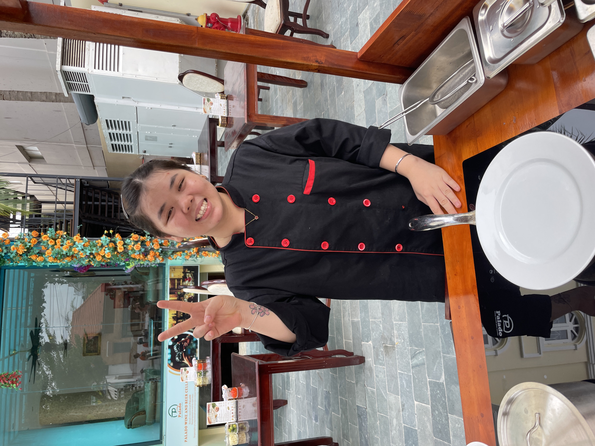 A smiling woman in a black chef's coat with red trim is photographed at Palado Wine and Beer Cellar in Phu Thuy Ward, Vietnam. She is making a peace sign with her left hand. She has a tattoo of flowers on her forearm. Next to her is a hot plate with a white frying pan. A long stainless steel container with a metal basket sits on the wooden counter nearby. The restaurant's glass window displays floral decorations and the restaurant's menu. The outdoor seating area is covered with patterned gray tiles. A large industrial air conditioning unit is visible on the building's exterior.