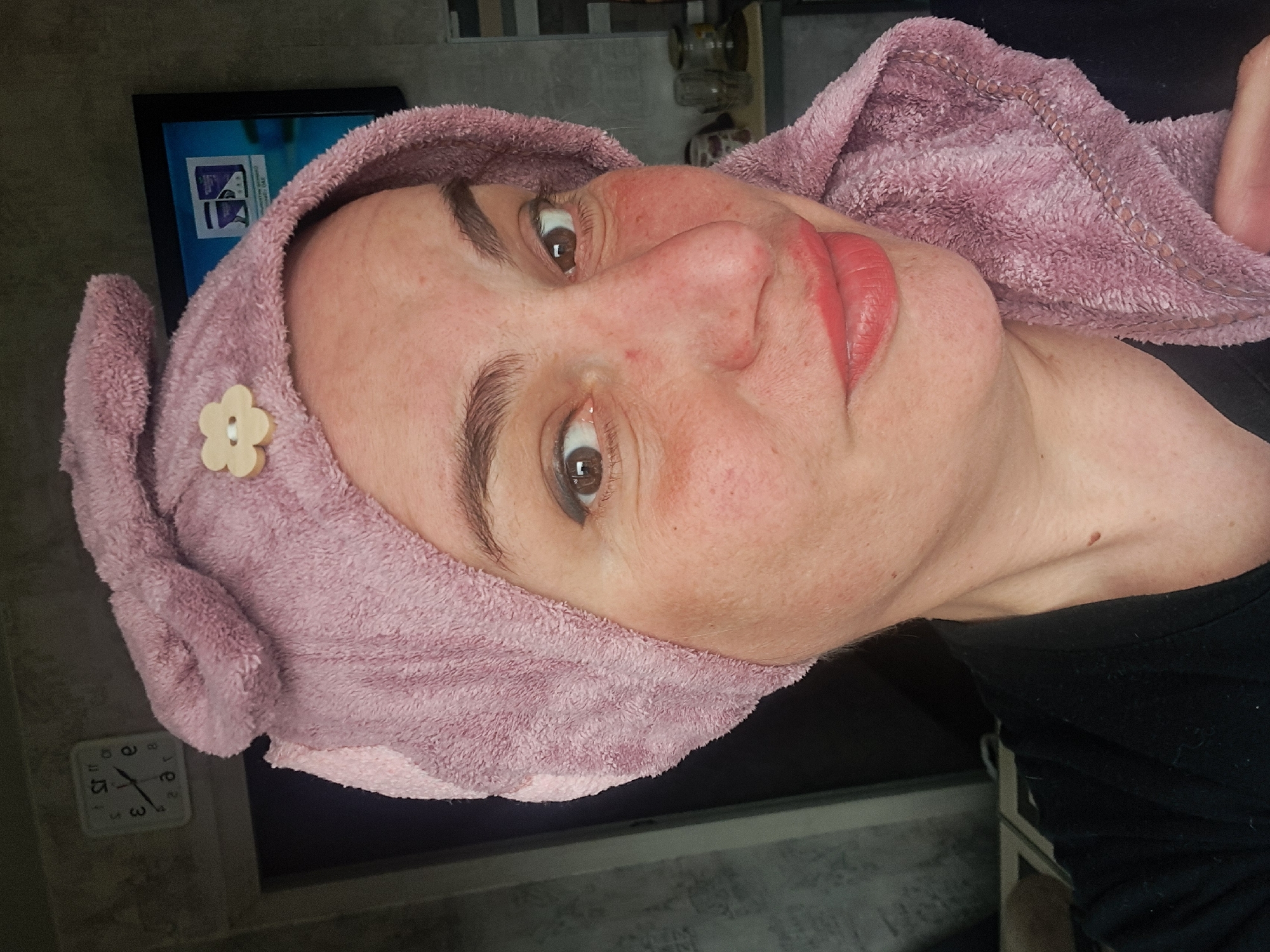 A medium close-up shot depicts a Caucasian woman with light skin. She has brown eyes and red lipstick. Her hair is wrapped in a pink towel adorned with a beige flower-shaped embellishment. She is wearing a black shirt. In the background, a white square clock is visible on a gray wall, and a framed screen displaying a blue image can be seen. The location appears to be indoors, but the city of Elektrostal, Russia, is not discernible from the image.