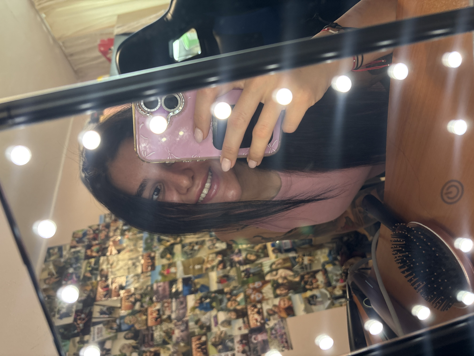A woman with dark hair and a pink shirt smiles while holding a pink phone in front of a vanity mirror with lights. The mirror reflects a wall covered in photos and a hairbrush. The woman is in Veliko Tarnovo, Bulgaria.
