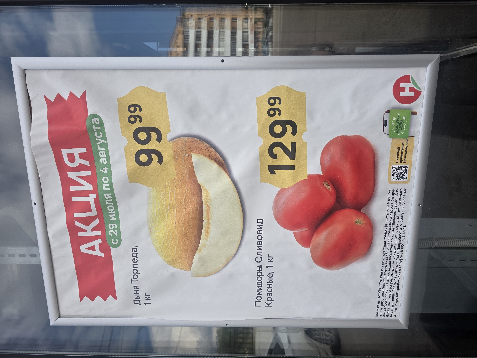 This is a photograph of a promotional poster displayed in a window, likely in a store. The poster advertises a sale on "Дыня Торпеда" (Torpedo Melon) and "Помидоры Сливовид Красные" (Plum-shaped Red Tomatoes). The melon is priced at 99.99 and the tomatoes at 129.99. The sale runs from July 29th to August 4th. The poster also includes QR codes and logos, one of which appears to be for a loyalty program. The background shows a reflection of a building and trees, indicating an outdoor setting. The poster is framed by a white border. No people are visible.