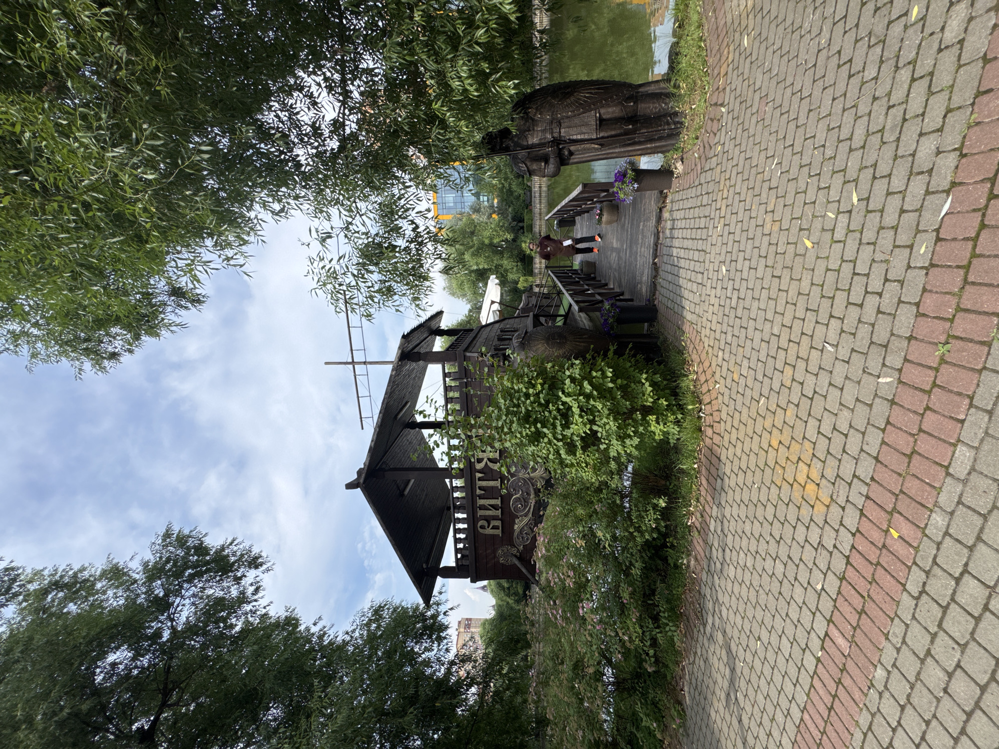 In Sovkhoz imeni Lenina, Russia, a wooden boat-shaped restaurant with Cyrillic lettering that reads "ВИТЯ" is situated on a body of water. A wooden walkway extends from a brick-paved path to the restaurant. A single individual, dressed in a brown shirt and dark pants, walks on the walkway. Two large, dark statues of figures in armor stand at the entrance to the walkway, flanking potted plants with purple flowers. Lush green trees and bushes surround the scene, and a cloudy sky is visible above. In the background, a modern building with a yellow-orange facade can be seen through the trees.