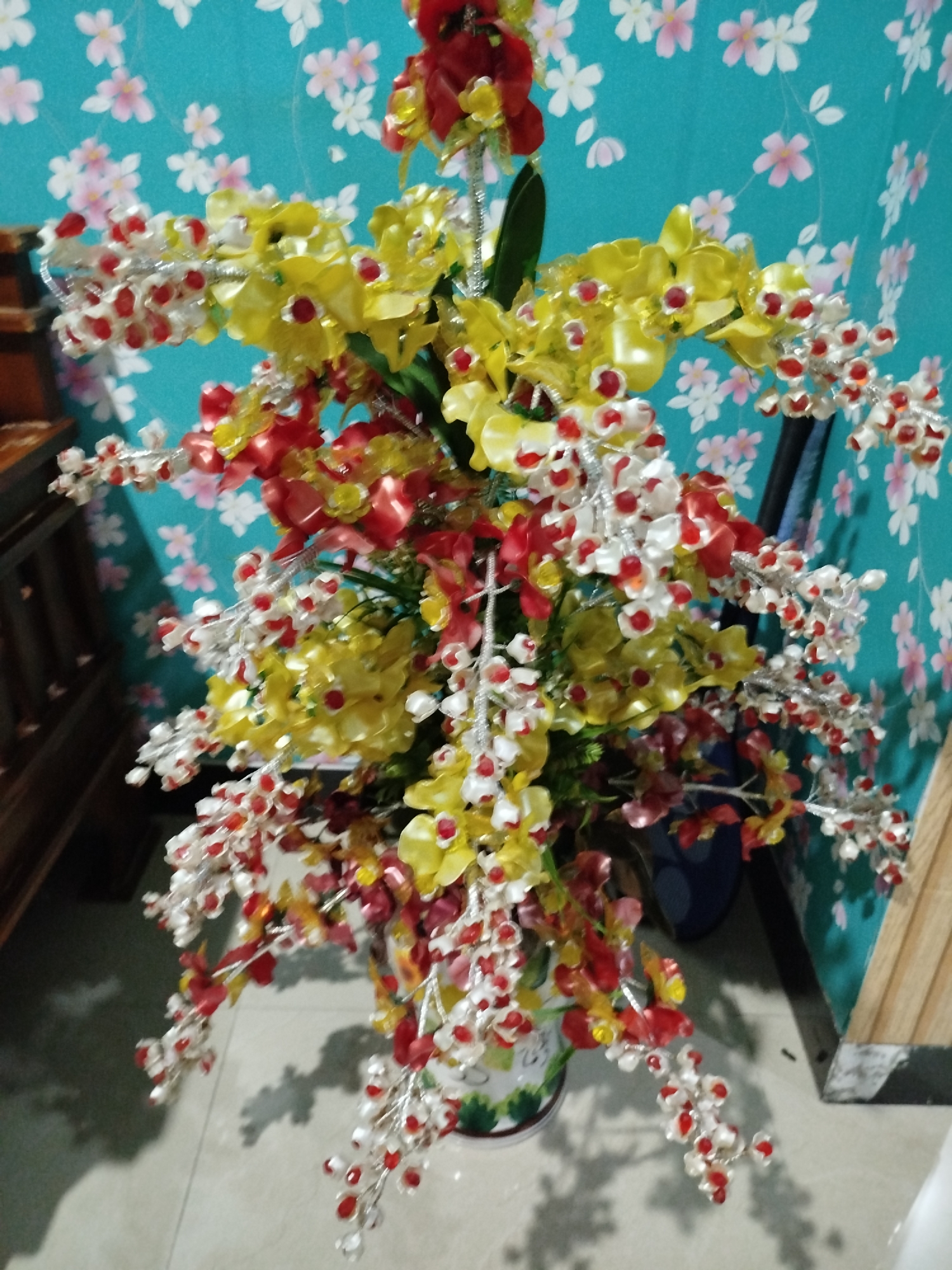 A close-up, indoor shot captures a vibrant artificial floral arrangement. The bouquet is dominated by bright yellow orchid-like flowers and numerous smaller clusters of white and red bead-like flowers attached to delicate silver stems. The arrangement is housed in a white vase adorned with green floral patterns. Behind the flowers, a teal-colored wall with a repeating pattern of pink and white flowers serves as the backdrop. To the left, a portion of a wooden piece of furniture is visible, and to the right, a wooden plank is partially seen. The scene appears to be set in a room in Bandungrejo, Indonesia.