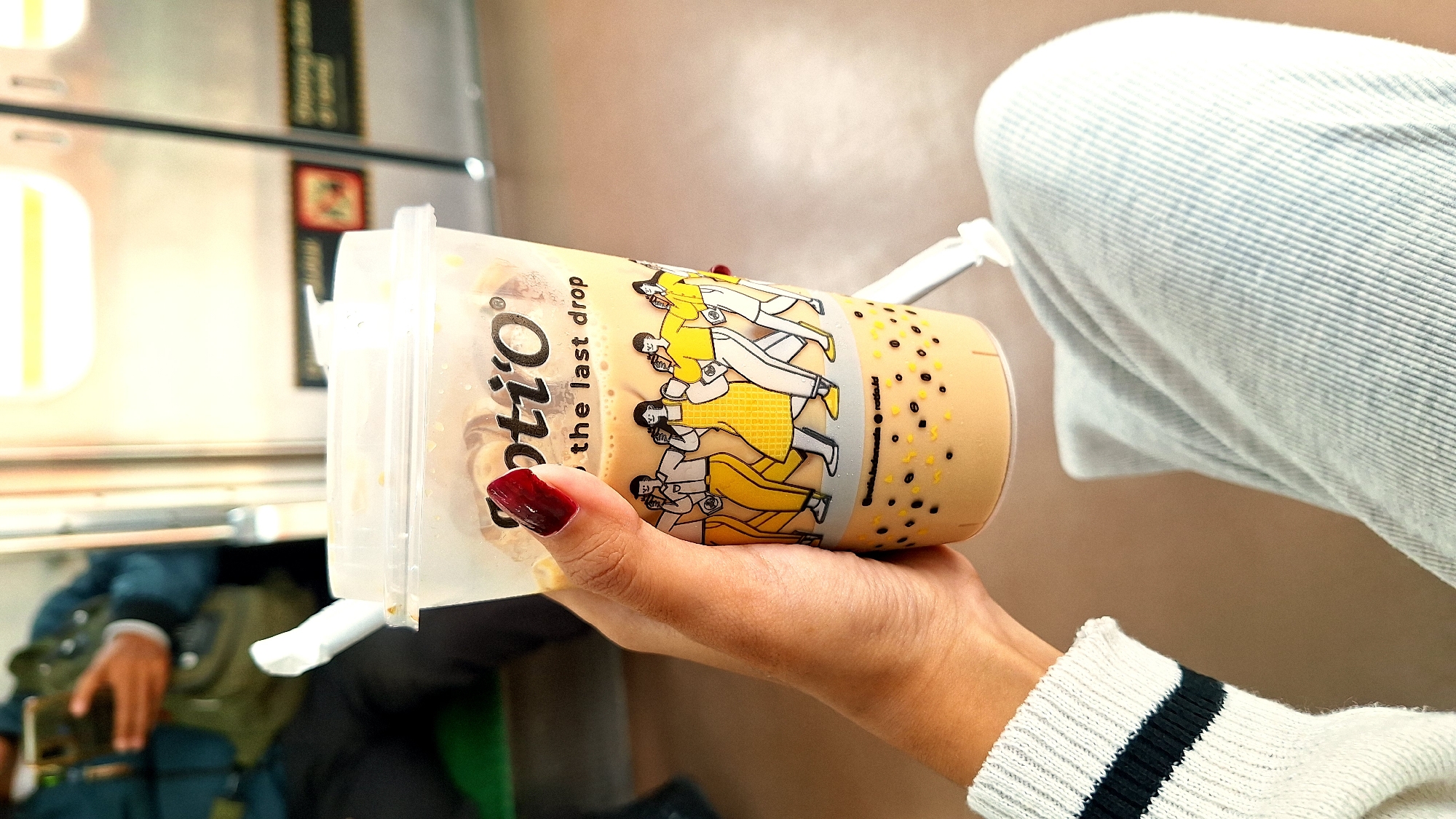 A photograph shows a person holding a plastic cup of bubble tea, identified by the logo "OTIO The Last Drop." The person's hand has dark red nail polish. They are wearing a white sweater with black stripes on the sleeve and light blue jeans. The background shows a train car interior, with an unidentified person partially visible in the lower left, wearing a dark blue jacket. Part of the interior includes metal paneling. The location, although not visually obvious, is Cikarang, Indonesia.
