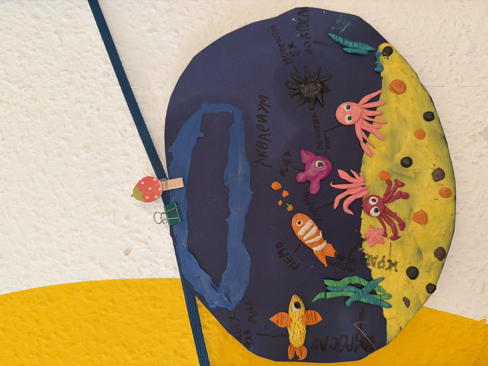 The image shows a colorful, handmade craft depicting an underwater scene. The craft is a dark blue, oval-shaped cutout with various clay figures attached. In the center, there's a depiction of an aquarium with a blue wavy line representing water. Various sea creatures made of clay are present, including a yellow fish, a clownfish, pink and purple octopuses, a pink starfish, and a black spider-like creature. The bottom of the craft is yellow, representing the seabed, with small black and orange circles that could be rocks or shells. There are also green clay sea plants. Text is written on the blue background, some in Russian, such as "АКВАРИУМ" (Aquarium). The craft is attached to a textured wall with a yellow and white section on the left, using a green binder clip and a wooden clip shaped like a strawberry. A dark blue rope runs diagonally across the top of the image. There is no discernible location in Yekaterinburg, Russia.