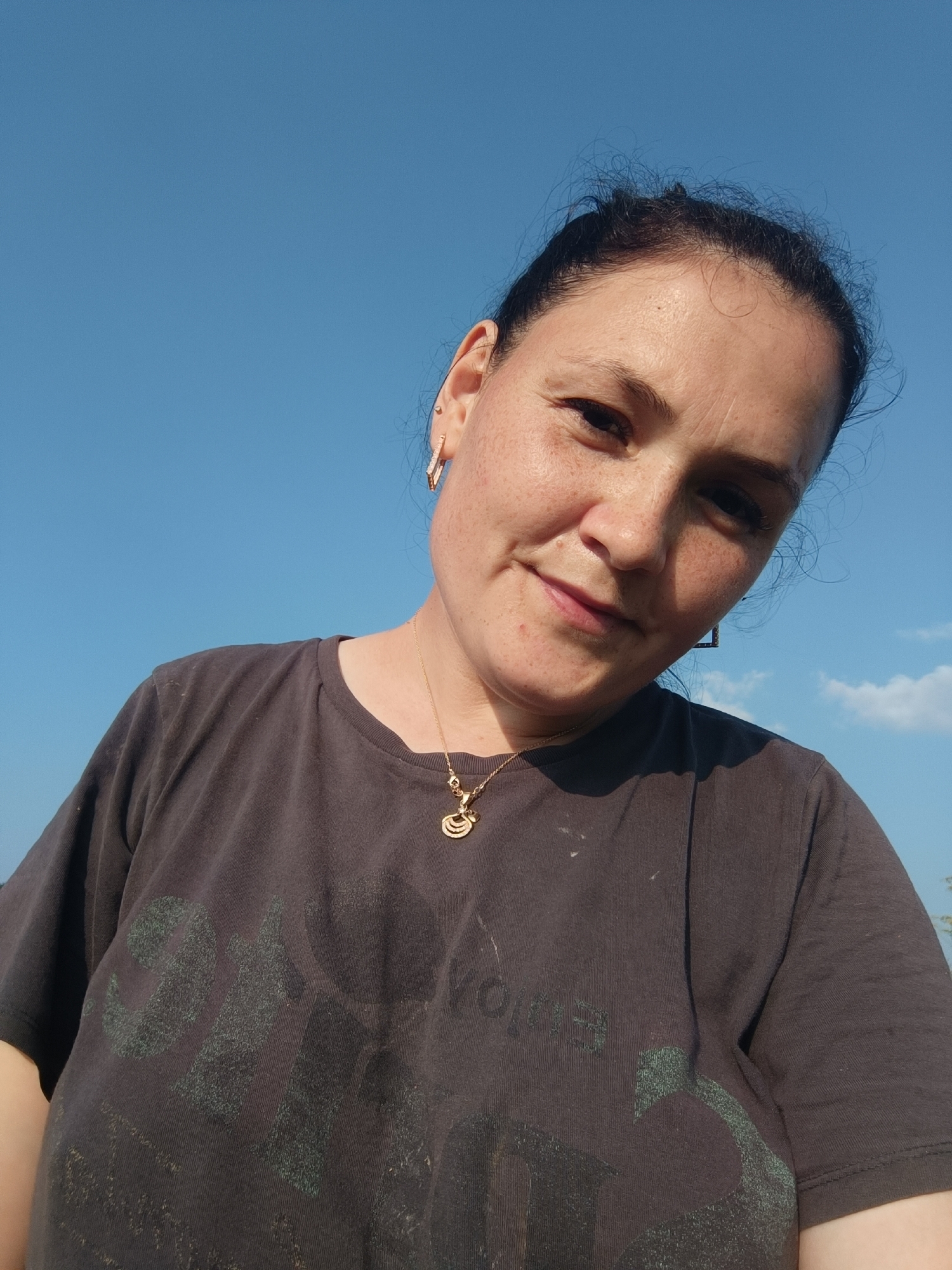 A woman is pictured outdoors against a clear blue sky. She is wearing a dark t-shirt with faded green text and a gold necklace with a pendant. Her hair is dark and pulled back, and she has freckles on her face. She is looking towards the camera with a slight smile. The image appears to be a selfie. There is no information about the location of Karsha, Russia in the image.