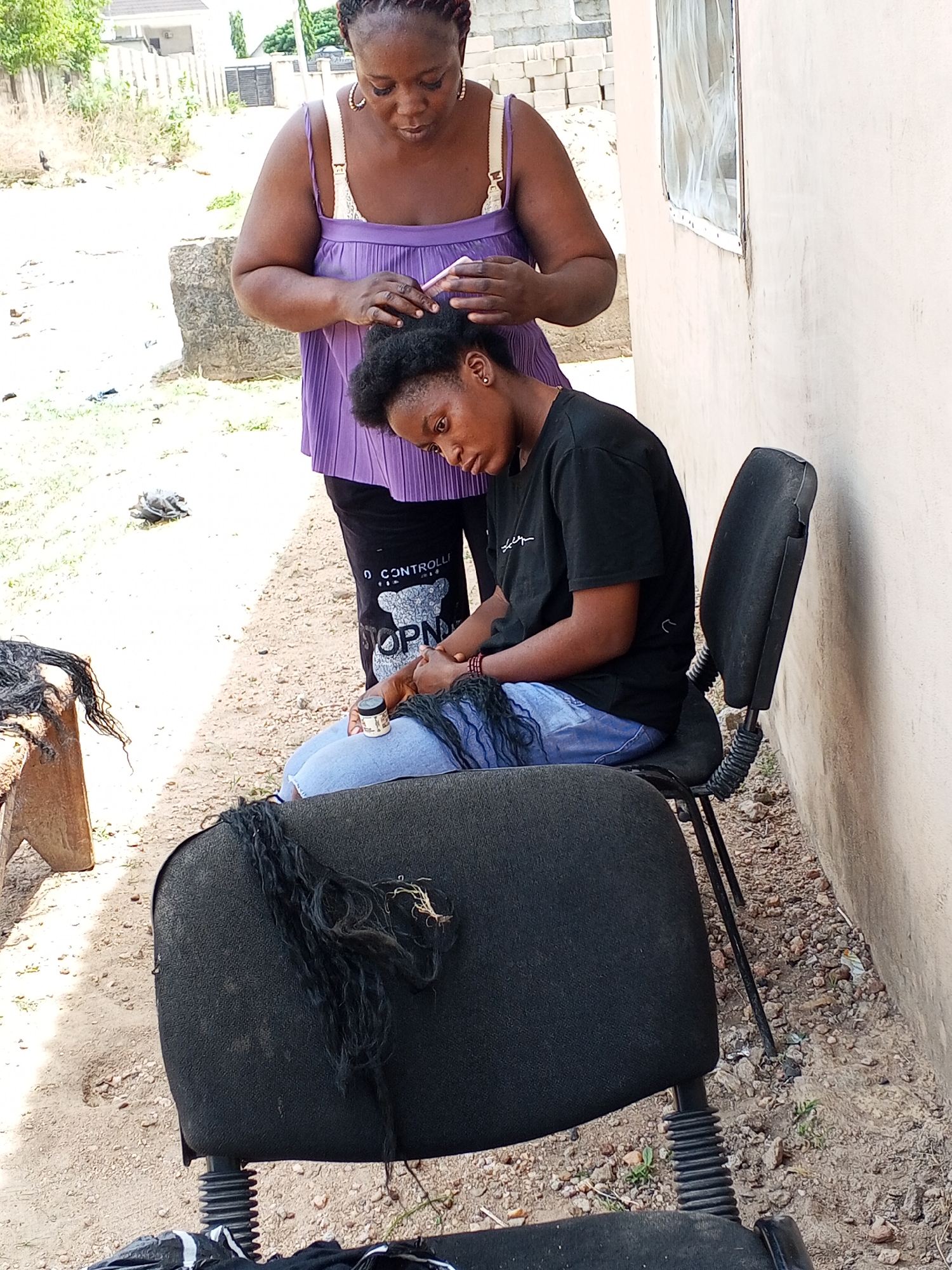 In Adonkolo, Nigeria, a bright outdoor scene captures two women engaged in hair styling. An older woman, with braided hair and hoop earrings, stands dressed in a purple pleated top and black pants, which bear partial text "0 CONTROLLED" and "TOPN." She is actively combing the hair of a younger woman, who is seated on a black office chair, wearing a black t-shirt with unreadable white text and light blue jeans.
The seated woman leans forward, holding a small white jar of hair product, seemingly applying it or preparing for the process. Black hair extensions are draped over her lap and on the chair's back. To the left, a wooden stool also holds a pile of black hair extensions. The setting features an unpaved, dusty ground with scattered small stones and sparse vegetation. A plain, light-colored building wall with a simple window and sheer white curtains forms the backdrop to the right. In the distance, other buildings, a gate, and trees are visible under clear, sunny daylight. The scene conveys a focused, routine activity in a casual, outdoor environment.