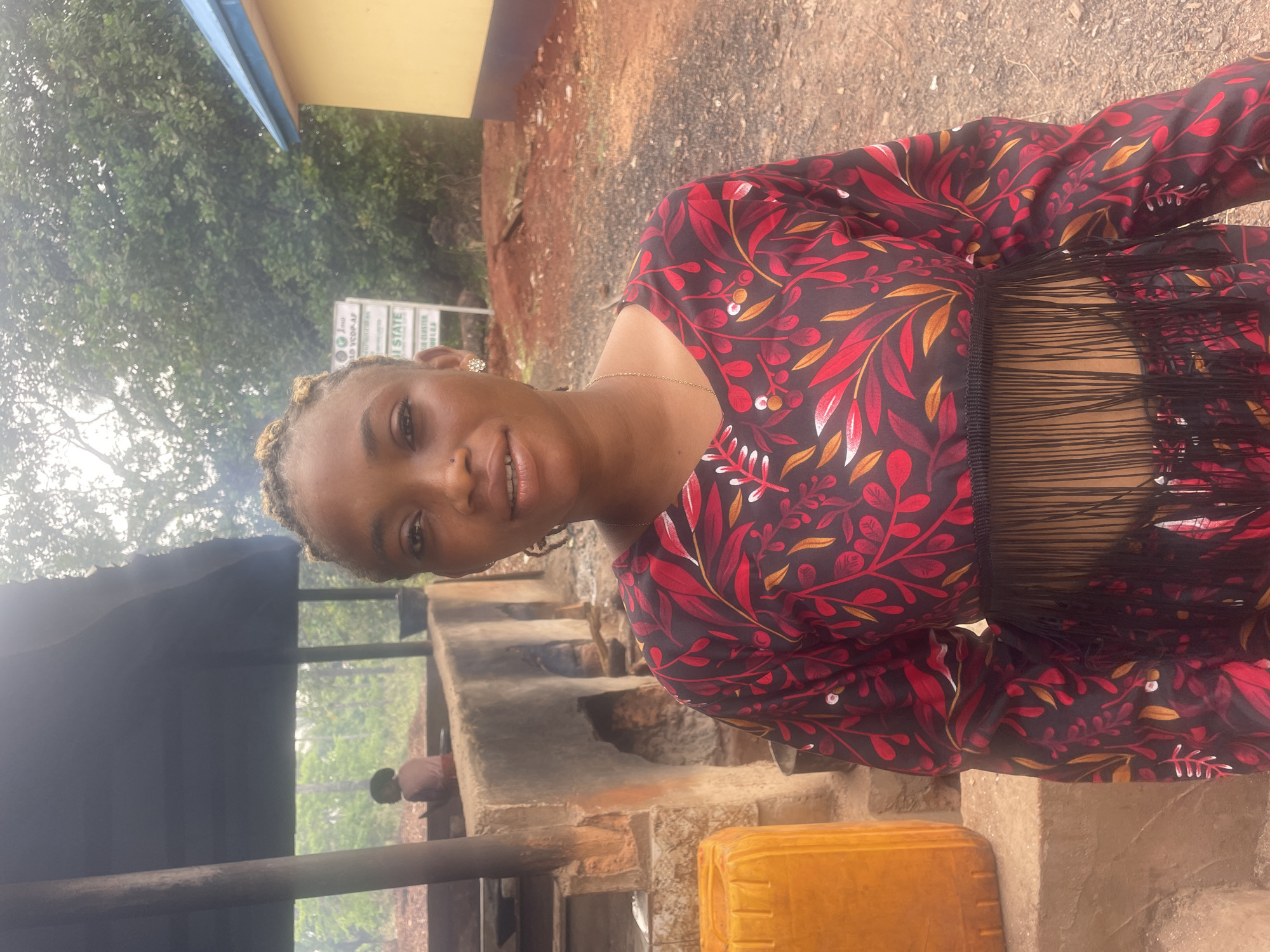 A woman with braided blonde hair stands outside in Eke, Nigeria, wearing a dark long-sleeved top with a red and gold leaf pattern and a fringe detail across the chest. She is smiling. Behind her is a cement structure with openings, possibly used for cooking, with what appears to be smoke rising from it. A yellow plastic container is visible near her feet. The ground is brown and covered in dirt. In the background, there are trees and a building with a sign.