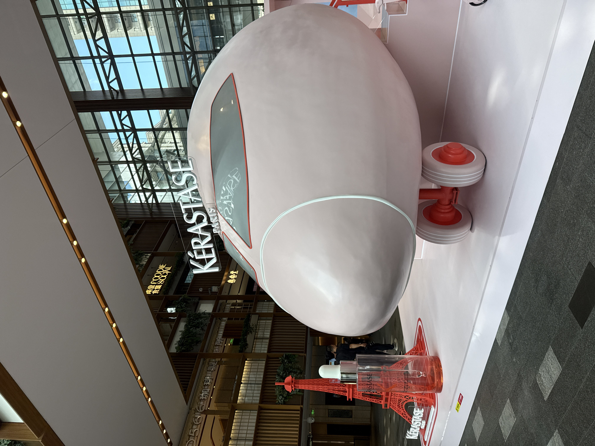 This is a photograph taken indoors, possibly in a shopping mall, in Shanghai, China. In the foreground, a large, light pink, airplane-shaped sculpture is the main subject. The sculpture has red accents around its windows and red wheels with white rims. To the left of the airplane sculpture is a miniature red replica of the Eiffel Tower. Next to the Eiffel Tower replica is a tall glass bottle containing orange liquid, with a dropper on top, which appears to be a Kérastase product. The Kérastase logo is visible on the bottle and on a sign behind the airplane sculpture. In the background, there are glass windows reflecting buildings and a bright blue sky. There are also signs visible, one of which reads "FOODIE SOCIAL". Several people are visible in the mid-ground, walking and standing. The floor is tiled with dark grey and lighter grey square tiles. The overall scene suggests a promotional display or event.