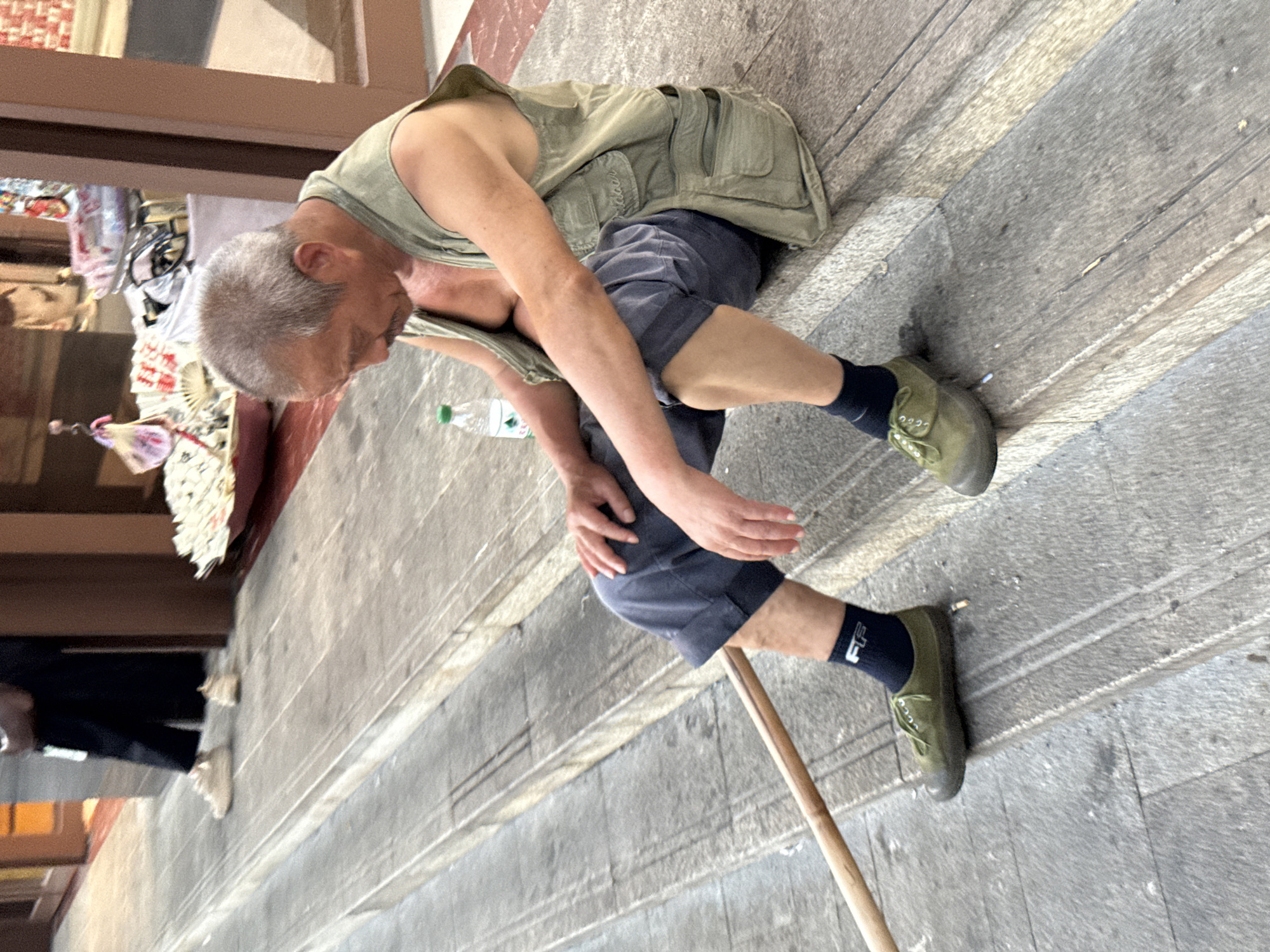 An older man with gray hair and a mustache sits on a concrete step, looking down. He wears a light green vest over a bare chest, dark blue shorts, black socks with a white logo, and olive-green canvas shoes. A wooden pole lies on the steps in front of him, and a plastic water bottle sits behind him. In the background, a person in dark pants and light-colored shoes stands near a doorway. Shelves with various items, including fans and what appear to be religious offerings, are visible in the background, suggesting a market or shrine area within Yuzhong District, China. The scene is set on outdoor stairs with a weathered appearance.