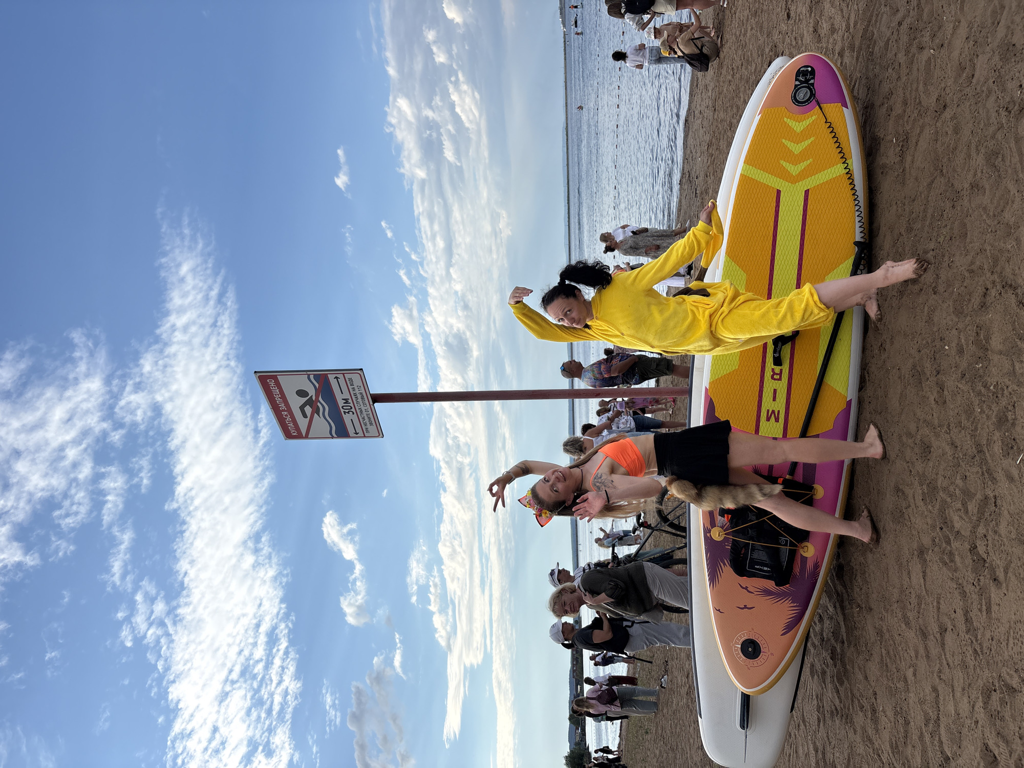The image depicts a group of people on a sandy beach next to a body of water. The scene appears to be a recreational area, likely a beach or shoreline. The sky is blue with scattered white clouds, suggesting it is daytime and the weather is pleasant.
In the foreground, two women are prominently featured. One woman, wearing a bright yellow jumpsuit, is posing on a paddleboard, with her arm raised in a flexing gesture. Her expression is energetic and playful. Beside her, another woman in an orange bikini top and black skirt is also posing on a paddleboard, making a peace sign with her fingers. She has a colorful headband and a furry tail accessory. Both women are barefoot and appear to be enjoying themselves.
Several other people are visible in the background, some standing and some sitting, suggesting a gathering or event. There is a sign in Russian that reads "КУПАТЬСЯ ЗАПРЕЩЕНО" which translates to "SWIMMING PROHIBITED". Below this, it indicates a distance of "30м" (30 meters) and instructions to call emergency services. The paddleboards themselves are colorful and decorated with designs. The overall atmosphere seems festive and relaxed, despite the prohibition sign. The presence of paddleboards and the beach setting indicate an outdoor water-related activity is likely occurring or has recently occurred. The location context provided is Chaikovsky, Russia.