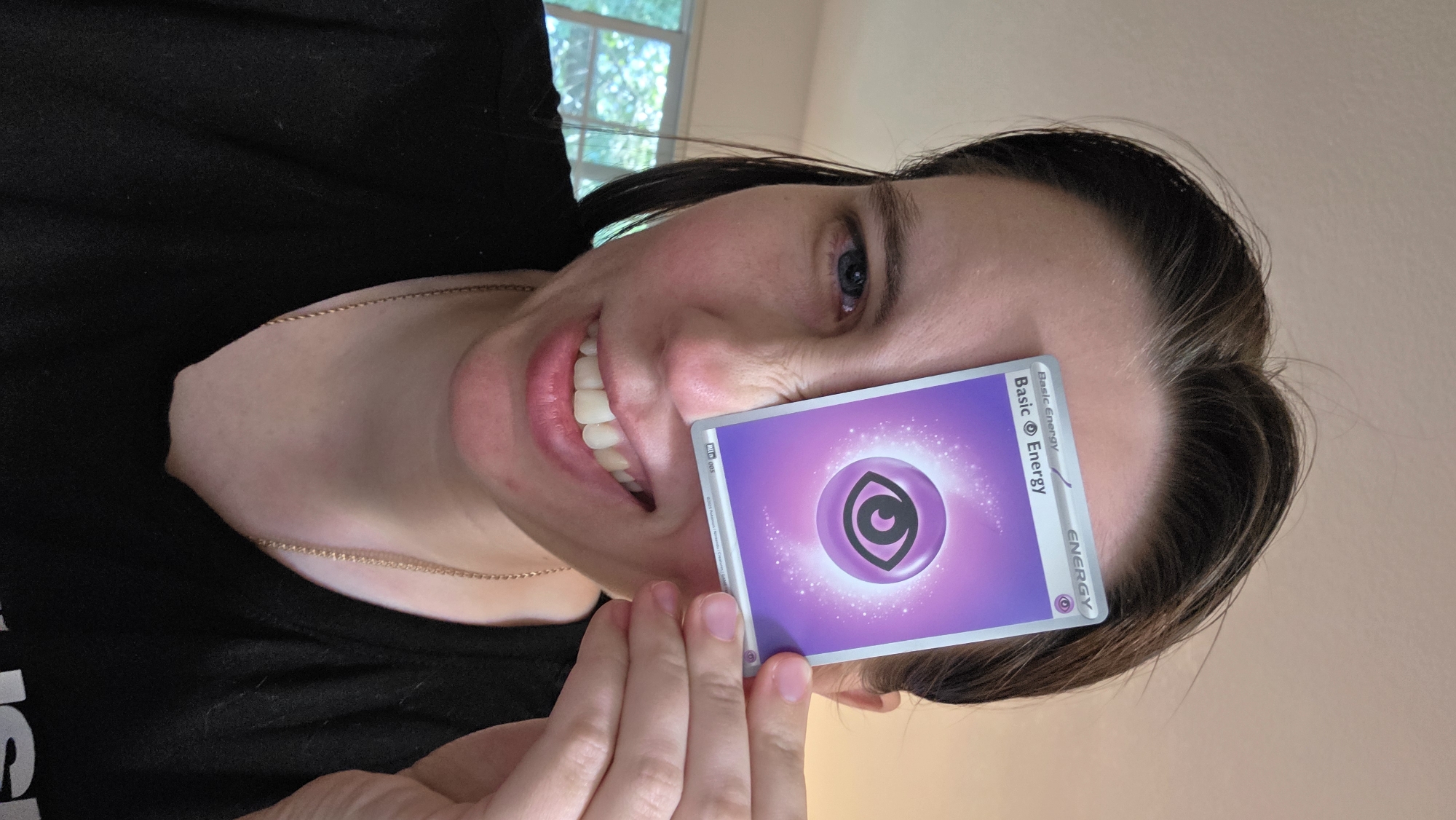In Portland, United States of America, a woman is captured in a close-up shot, holding a Pokémon card in front of her left eye, partially obscuring it. She is wearing a black t-shirt and a gold chain necklace. Her right eye is visible, and she is smiling, revealing her teeth. The background is blurred, with a hint of greenery suggesting an outdoor or indoor setting with windows. The lighting appears to be natural, possibly from a window or overhead light source.