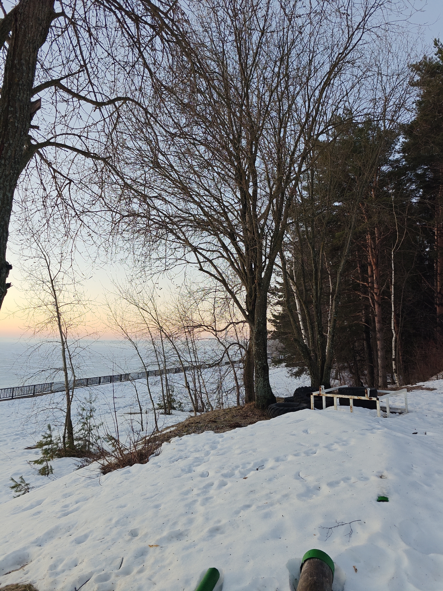 A winter scene in what appears to be the city of Chaikovsky, Russia. Snow covers the ground, and bare trees dominate the landscape. In the distance, a body of water is visible with a fence or walkway along the edge. Closer to the viewer, there's a pile of tires next to a white metal frame. The sky shows colors of dawn or dusk, with hues of pink and orange. Some green equipment is lying on the snow in the foreground. The scene is quiet and devoid of people.