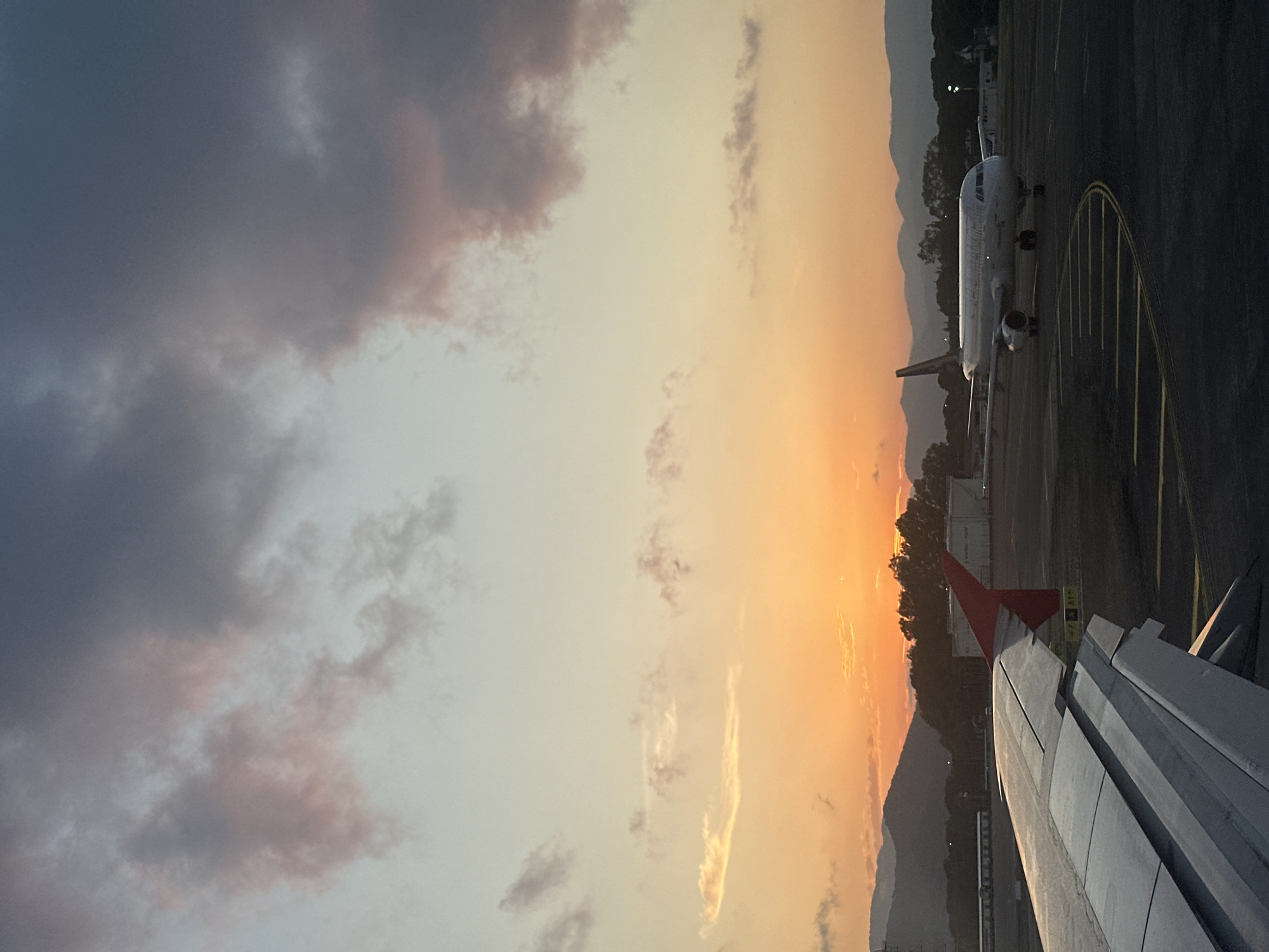 The image depicts an airport scene at sunset, observed from the window of an aircraft, presumably at Dalaman Airport in Dalaman, Turkey. The sky is dominated by a vibrant gradient of colors, transitioning from deep orange, red, and yellow at the horizon, where the sun is setting, to lighter blue and grey-pink in the upper portions. Scattered clouds, some tinged with pink and purple, are present across the sky.
In the lower left quadrant, the silver-grey wing of the observer's airplane is prominently featured, displaying its flaps and a red wingtip fence. The wing is sharply detailed, with visible panel lines and structural elements.
In the midground, on the tarmac to the right, another white narrow-body passenger aircraft, likely an Airbus A320 series, is stationary. It is oriented with its nose slightly turned towards the viewer, revealing its cockpit windows and two underwing engines. Yellow painted lines, indicative of taxiways or parking positions, are visible on the dark grey asphalt tarmac around this aircraft.
The background features a range of dark, silhouetted mountains stretching across the horizon. Below the mountains, obscured by the low light, are dark trees and various airport infrastructure elements, including what appear to be industrial tanks and buildings. No individuals are visible in the scene. The overall lighting suggests the transition from late afternoon to evening.