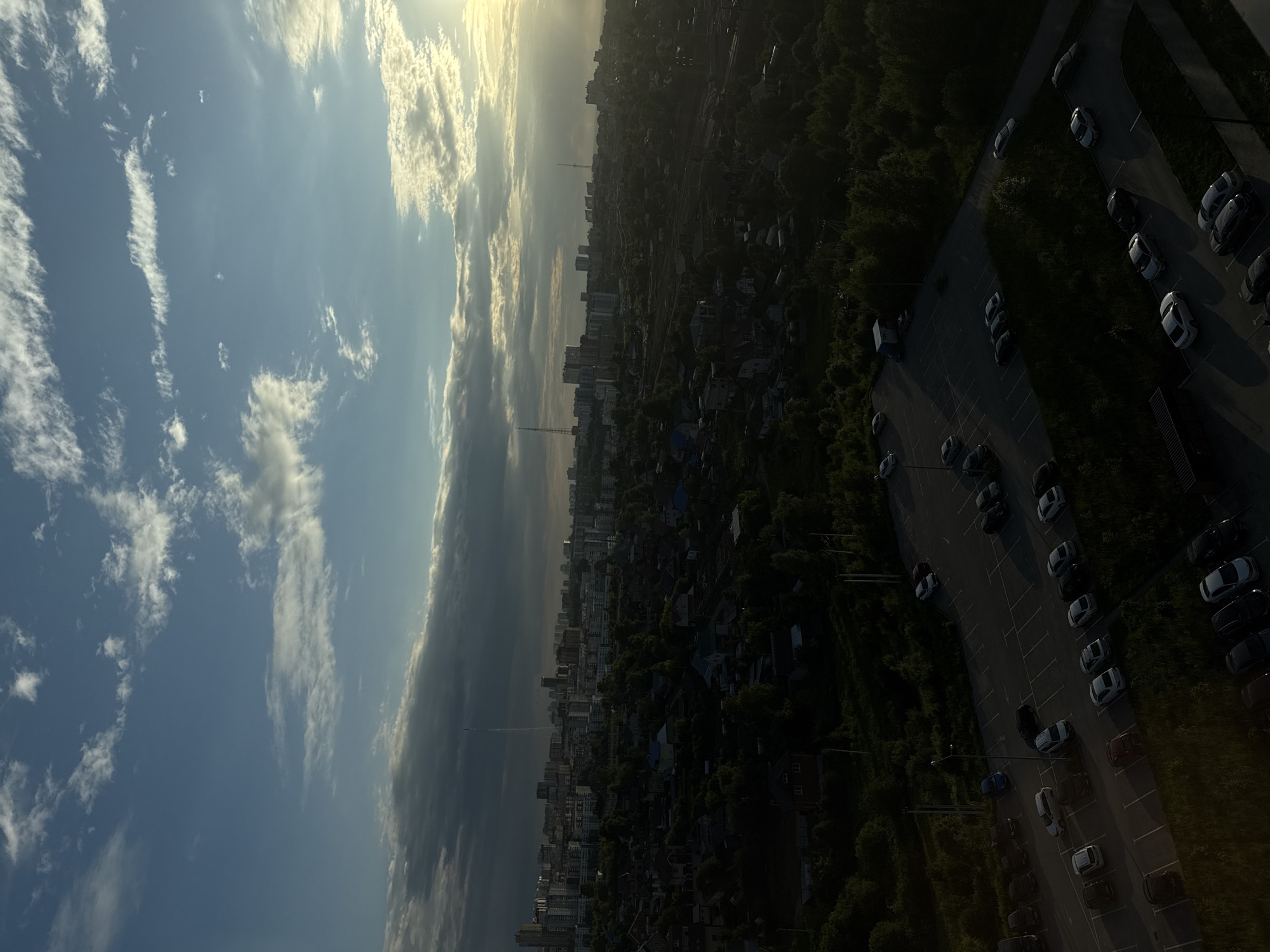 This is a high-angle, long shot aerial photograph of a city skyline and a parking lot in Perm, Russia. The sky is partly cloudy with a sunset casting a warm glow on the scene. In the distance, a dense urban area with various buildings, including high-rises and smaller structures, is visible. A prominent television tower stands tall among the buildings. In the foreground, a large parking lot filled with numerous cars is depicted, bordered by lush green trees and vegetation. The lighting suggests it is late afternoon or early evening.