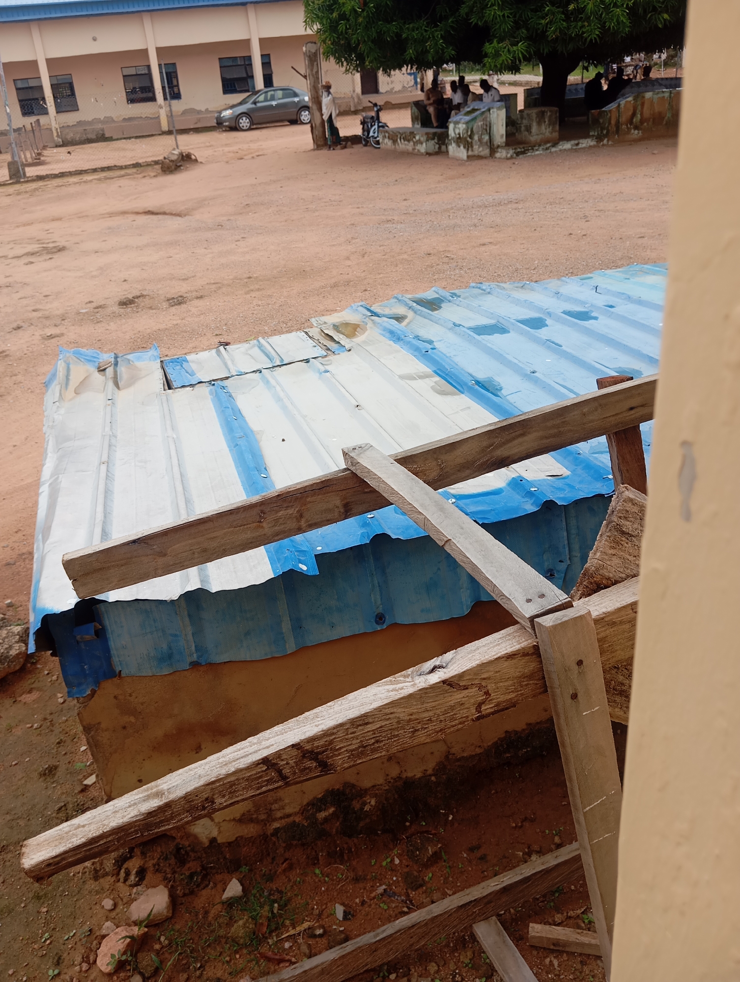The image depicts an outdoor scene in Jalingo, Nigeria, focusing on a dilapidated corrugated metal structure with blue and white sheeting. The structure appears to be a makeshift shelter, possibly a stall or an extension, with visible signs of disrepair and some wooden supports. In the background, a light-colored building with multiple windows is visible, suggesting it might be a school or administrative facility. A grey sedan is parked in front of the building. Further in the background, under the shade of a large tree, a group of people are seated, engaged in conversation or activity. A bicycle is also present near the tree. The ground is predominantly red dirt, common in many parts of Nigeria, with some scattered rocks and sparse vegetation. The overall impression is one of a functional, possibly semi-urban or peri-urban environment with a mix of permanent structures and more temporary constructions.