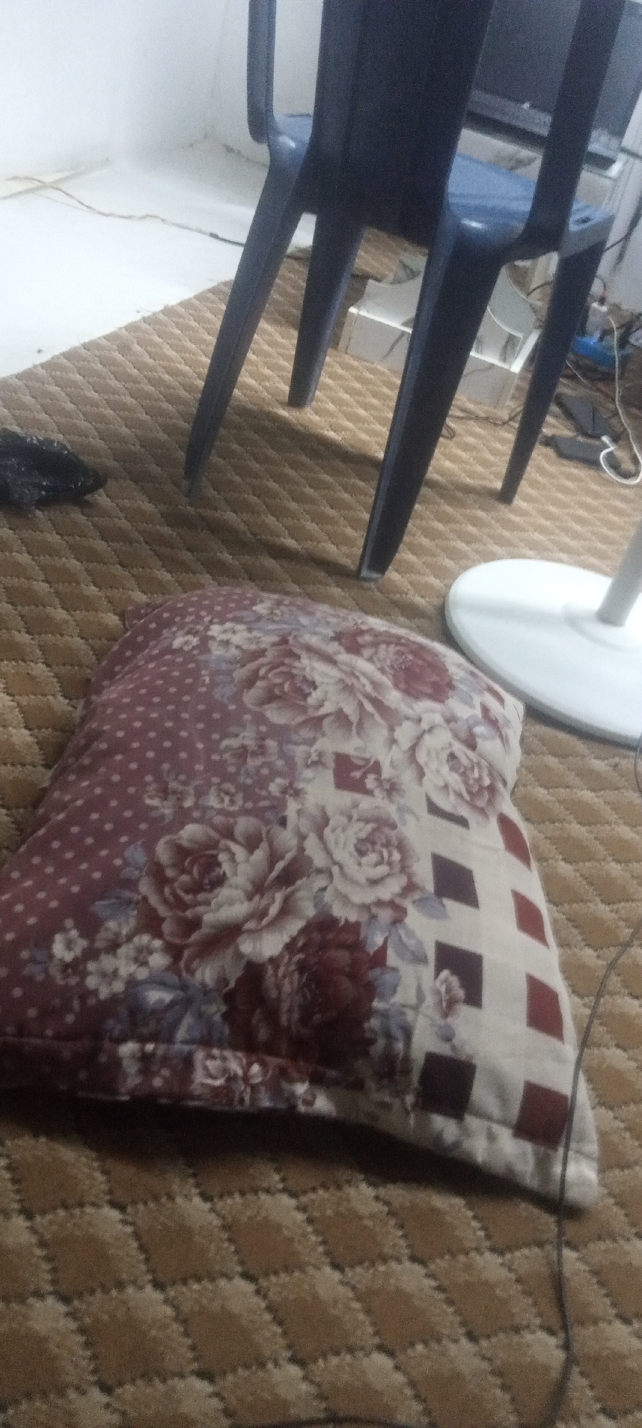 A medium shot shows a pillow on a textured rug, near a chair and a fan in an indoor setting. The pillow has a floral and geometric pattern in maroon, pink, and white tones. It rests on a brown rug with a small, repeating diamond pattern. To the upper right is a blue plastic chair with a visible section of a white fan base. A black object is slightly blurred and off to the left of the pillow. The environment suggests an interior space, likely a room. The location is Unknown, Unknown.