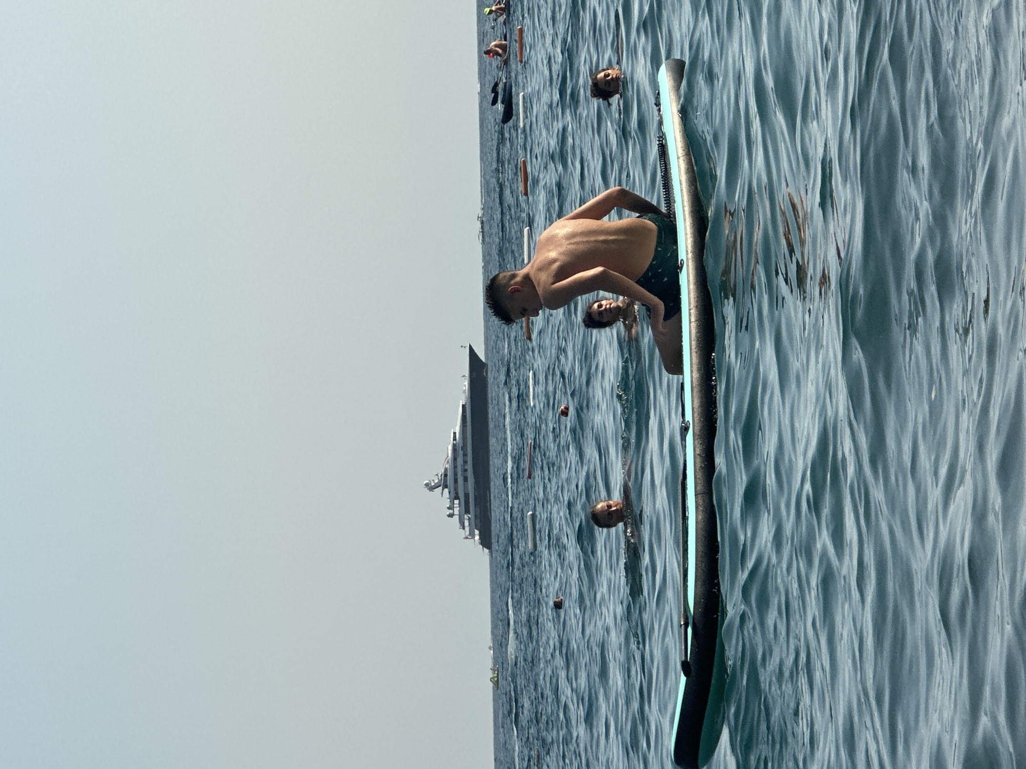 The image depicts a clear, sunny day at sea, likely in the vicinity of Boreti, Montenegro. In the foreground, a young person with dark hair and wearing dark swim trunks sits on the edge of a blue and black paddleboard, facing away from the camera. The paddleboard is partially submerged in the choppy blue water. In the mid-ground, several other individuals are in the water. To the left, two people are partially submerged, looking towards the camera. To the right, another person is also partially visible in the water. Further in the background, a large, multi-decked yacht with a dark hull and white superstructure is anchored on the horizon. Several buoys or markers are visible on the water's surface. The sky is a pale, hazy blue, suggesting a warm and clear day. The overall scene conveys a sense of leisure and recreation on the water.