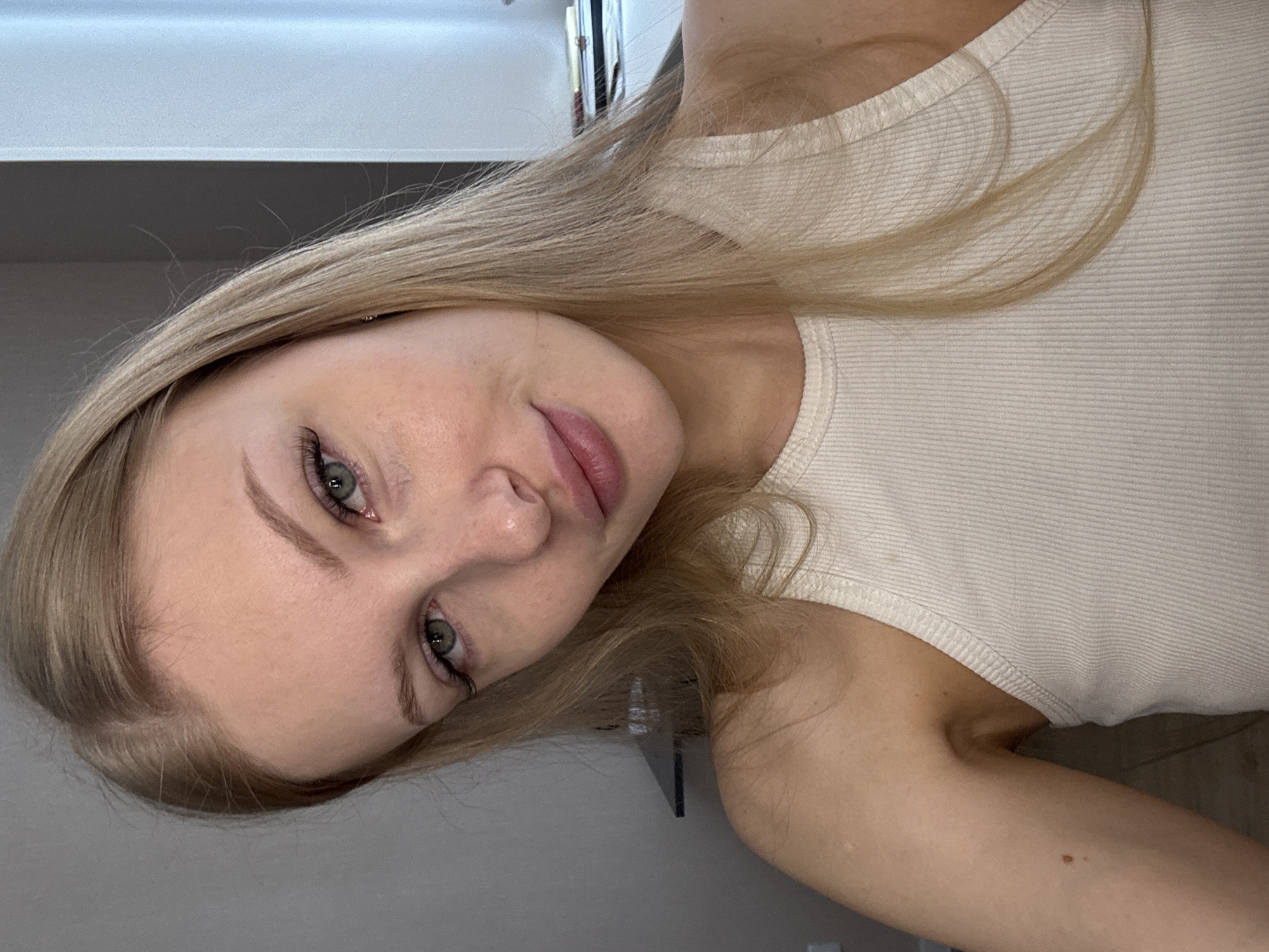 A young woman with blonde hair and green eyes is looking at the camera. She is wearing a beige tank top. Her makeup includes mascara and lip gloss. Her skin has a few freckles. The background is blurry and appears to be an indoor setting. There is a hint of natural light coming from the right side of the frame. The overall impression is that of a casual, candid portrait. There are no discernible landmarks or elements that would indicate the location as Perm, Russia.