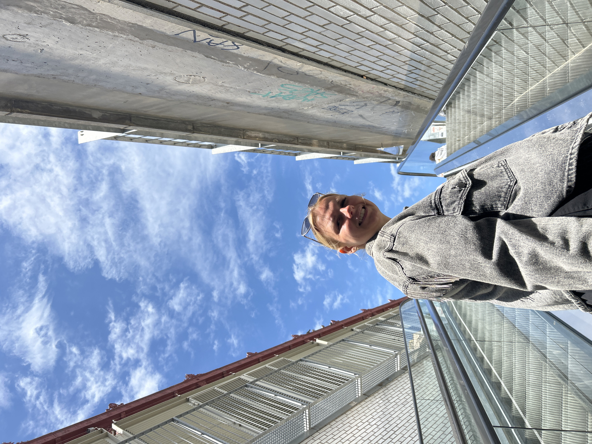 The image shows a person looking up at the sky while standing on a pedestrian bridge in Barcelona, Spain. The perspective is tilted upwards, capturing the person's face and upper body, as well as the surrounding architecture and sky.
The person is wearing a gray denim jacket and sunglasses are perched on their head. They are smiling and looking directly at the camera.
Above the person is the underside of a concrete bridge, which is marked with graffiti.
The sky is blue with scattered white clouds.
Below the person is a building with a brick facade and a red tile roof. The building has metal security grilles over its windows. A glass-enclosed pedestrian bridge is to the right of the building.