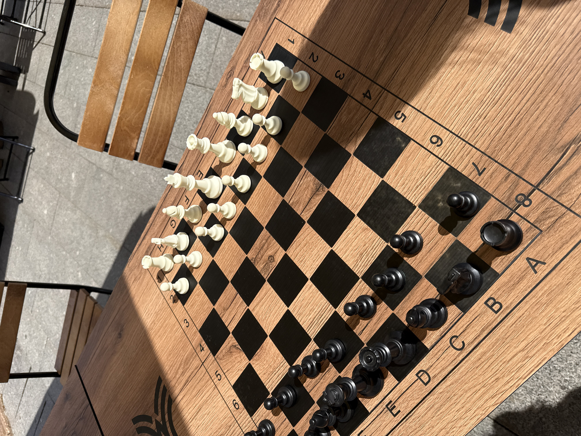 An overhead shot captures a wooden chess board set up outdoors, likely on a patio or in a park in Moscow, Russia. The board, which appears to be a table, features a light brown wood grain finish with contrasting black squares. All white and black chess pieces are arranged in their standard starting positions, suggesting a game is about to begin or has just been reset. The white pieces, made of a light-colored material, occupy the top-left portion of the board, while the black pieces, made of a dark material, are positioned on the bottom-right side.
Strong sunlight casts distinct, diagonal shadows across portions of the board and pieces, indicating a bright daytime setting, possibly morning or late afternoon. A slatted wooden chair with a black metal frame is partially visible on the left side, suggesting seating for players. The ground beneath appears to be paved with light gray tiles or concrete.
Visible alphanumeric coordinates are printed along the edges of the board. The numbers '1' through '8' are discernible along the left edge of the board, with '1' at the top (near the white pieces) and '8' at the bottom (near the black pieces). The letters 'A' through 'H' are visible along the bottom edge of the board, with 'A' on the right (near the black queen's rook) and 'H' on the left (near the black king's rook). A small, stylized black logo or emblem is faintly visible on the wooden surface in the bottom-left corner of the table.