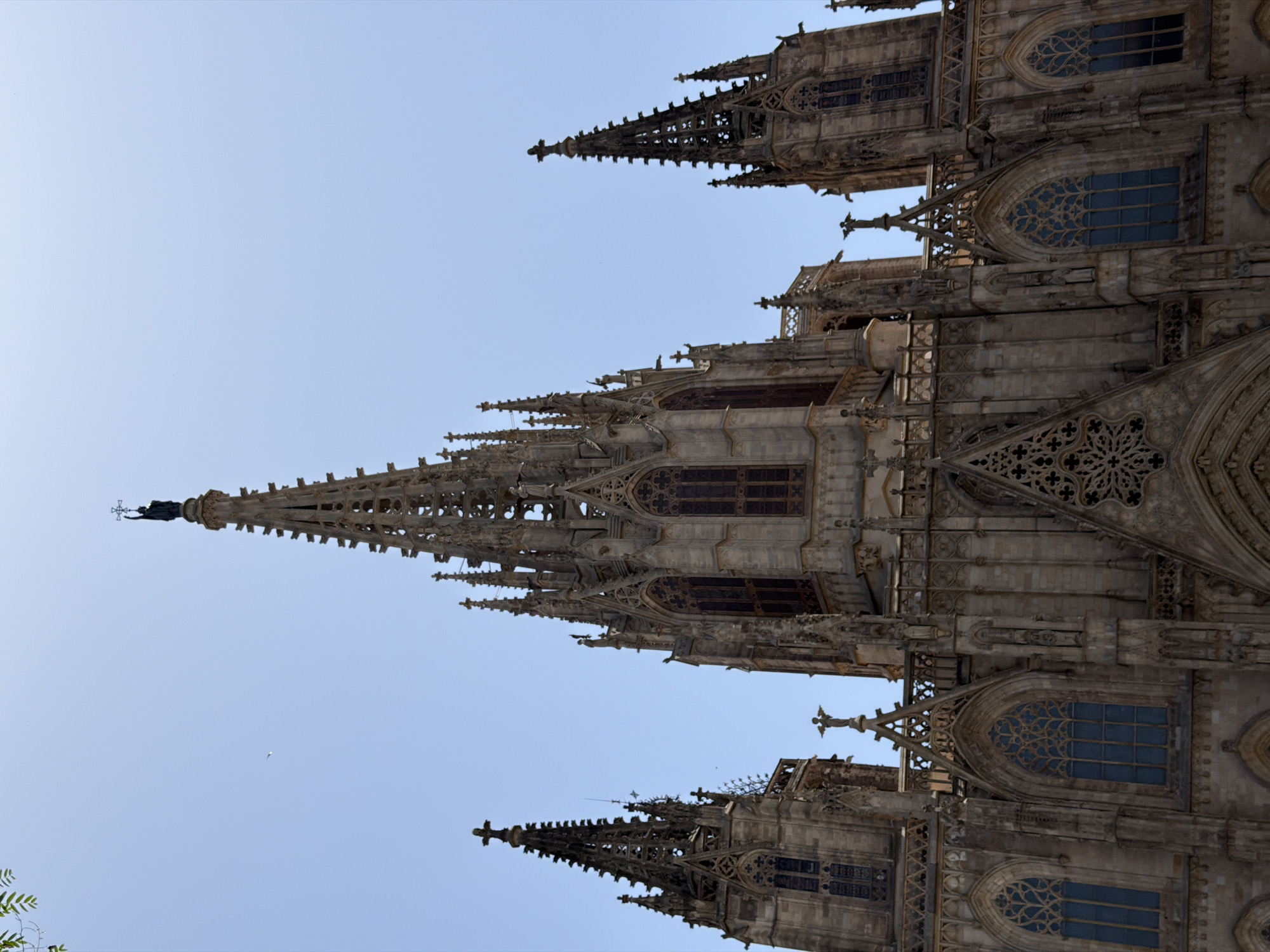 This is a low-angle, full-shot photograph of the Barcelona Cathedral, officially known as the Cathedral of the Holy Cross and Saint Eulalia, in Barcelona, Spain. The image captures the ornate Gothic architecture of the cathedral's facade, with its intricate stonework, towering spires, and numerous windows. The sky is a clear, bright blue, with a few faint birds visible. The cathedral is made of weathered stone, showing signs of age and exposure to the elements. The central spire is the tallest and most prominent feature, topped with a statue of a winged figure holding a cross. On either side of the central structure are two smaller spires, also elaborately decorated. The lower portion of the facade features arched windows with stained glass, some of which appear to be dark blue. The building is bathed in sunlight, casting shadows that accentuate the architectural details. There are no people or any other objects or elements present in the scene besides the cathedral and the sky.