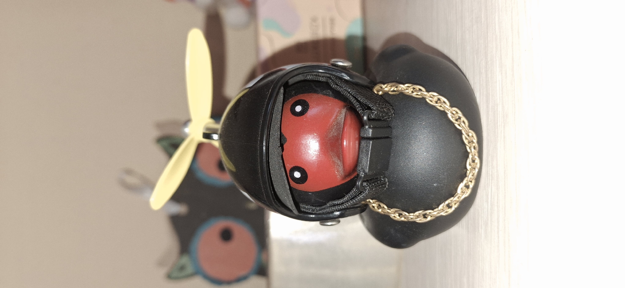 A close-up shot shows a car air freshener styled to resemble a ladybug, with a yellow propeller attached. The ladybug's body is red with white-bordered black eyes. It sits within a black, helmet-like frame, which is secured with black strapping. A gold-colored chain dangles from the side. The background appears to be a shelf in a room, with indistinct items behind the air freshener. The location, Писарёвка, Russia, cannot be verified with visual evidence.
