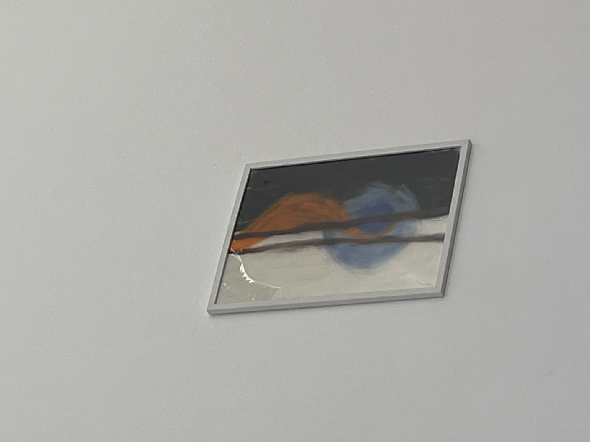 The image shows a framed abstract painting hanging on a white wall. The painting features blocks of black, white, orange, and blue paint. Two dark horizontal lines bisect the painting. The location is not evident from the image. The image is otherwise devoid of information about the city of Zlatograd, Bulgaria.