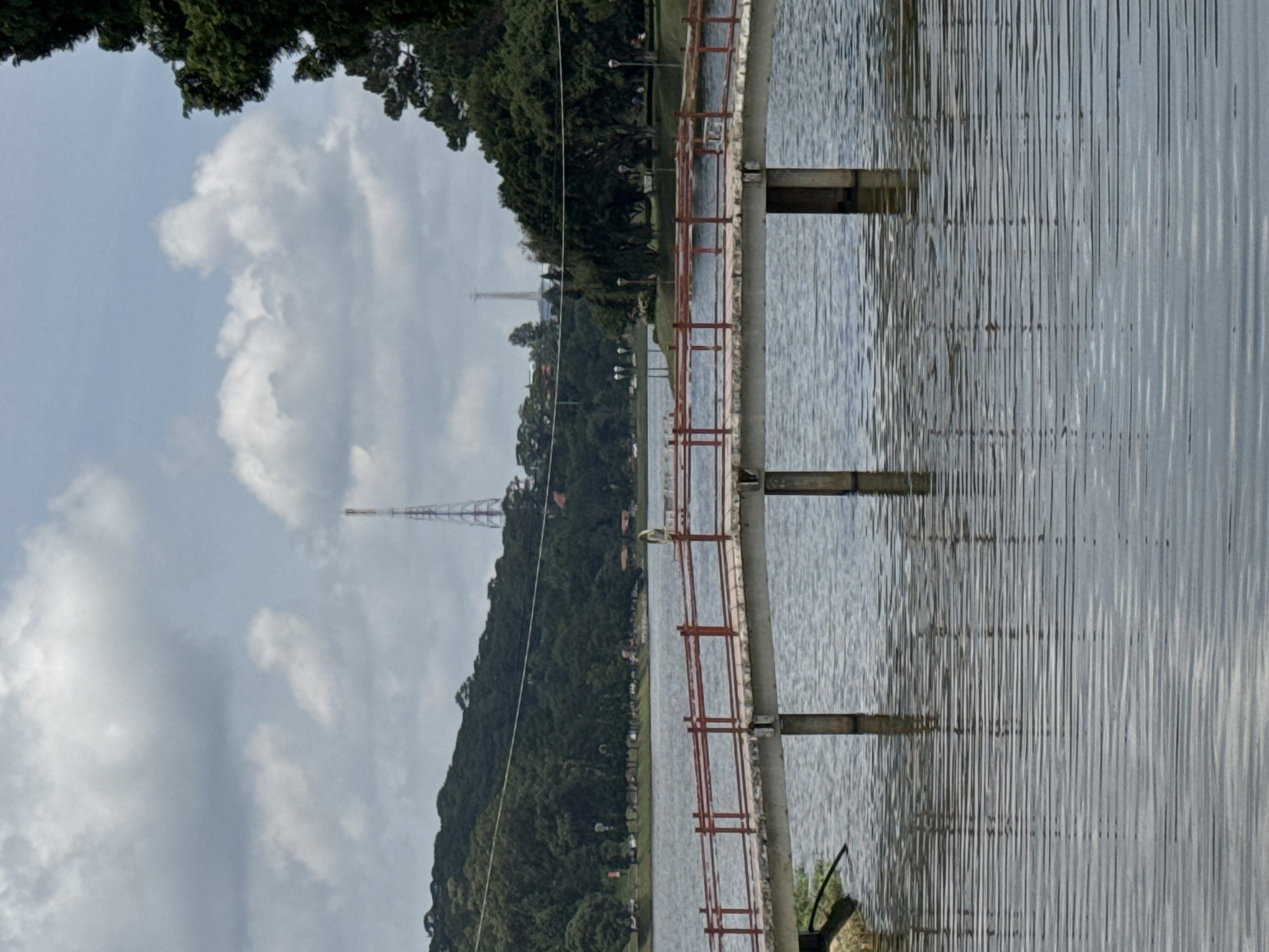 The image depicts Xuan Huong Lake in Da Lat, Vietnam, on an overcast day. A footbridge with red railings spans the water. The lake is calm, with visible ripples reflecting the sky and surrounding landscape. On the far side of the lake, a densely wooded hill rises, dotted with buildings and topped by a prominent telecommunications tower. The trees on the hill are lush and green. The sky is filled with a mix of white and gray clouds, suggesting a cloudy or potentially rainy atmosphere. No people are clearly visible on the bridge or in the immediate vicinity. The overall scene conveys a tranquil and natural setting within an urban environment.
