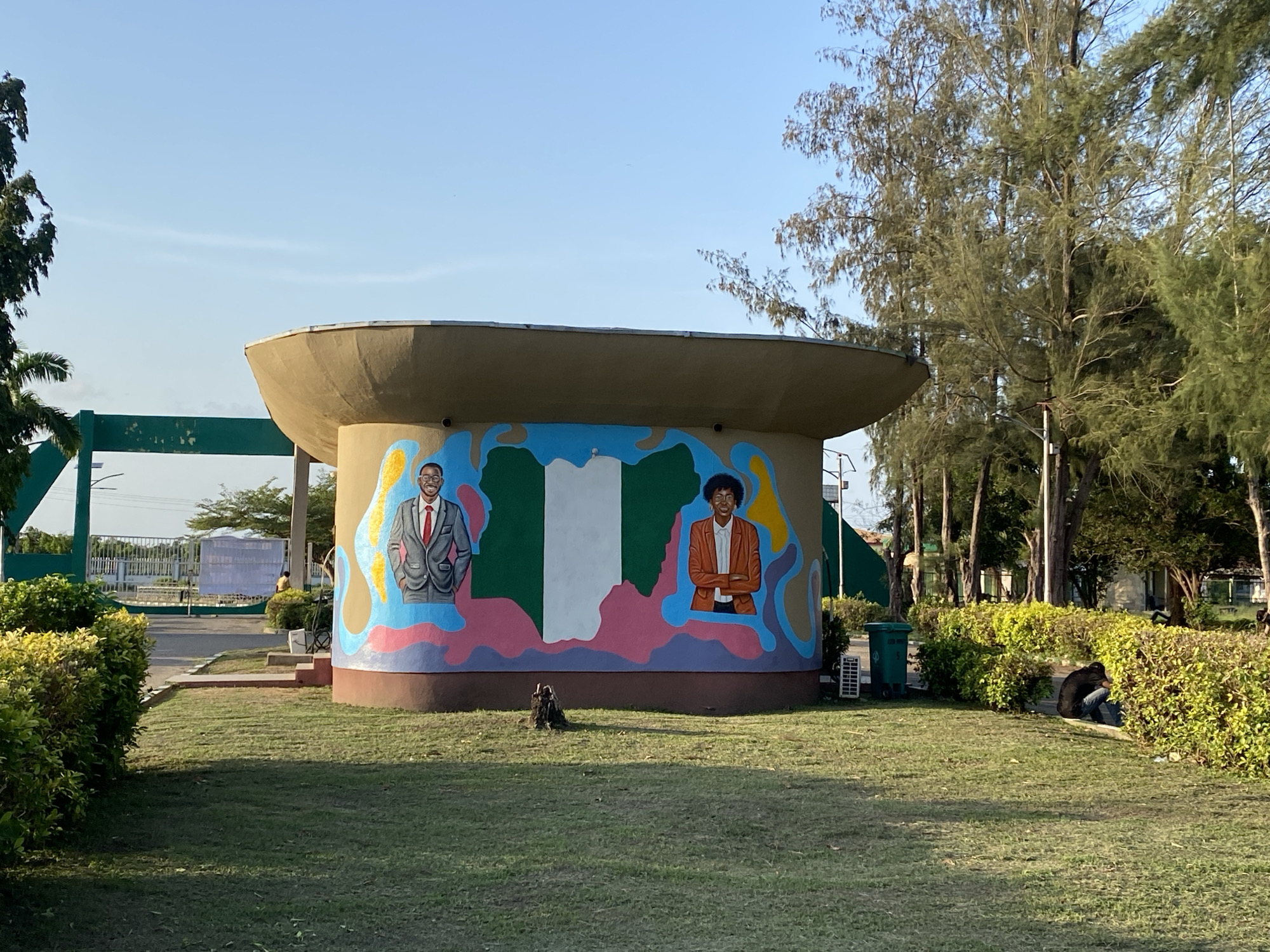 The image shows a circular, dome-like structure with a mural depicting the map of Nigeria and portraits of two individuals in Tawpo, Nigeria. The mural is painted on the side of the structure. The Nigerian flag forms the center of the map. To the left of the map is a man in a gray suit with a red tie, and to the right is a woman in a brown suit. The structure is on a grassy area, with bushes and trees in the background. A person is sitting near the bushes to the right of the structure. There is a green trash can next to the structure. In the far background is a green and white gate structure. The sky is blue and clear. A dark brown stump is present near the base of the structure.