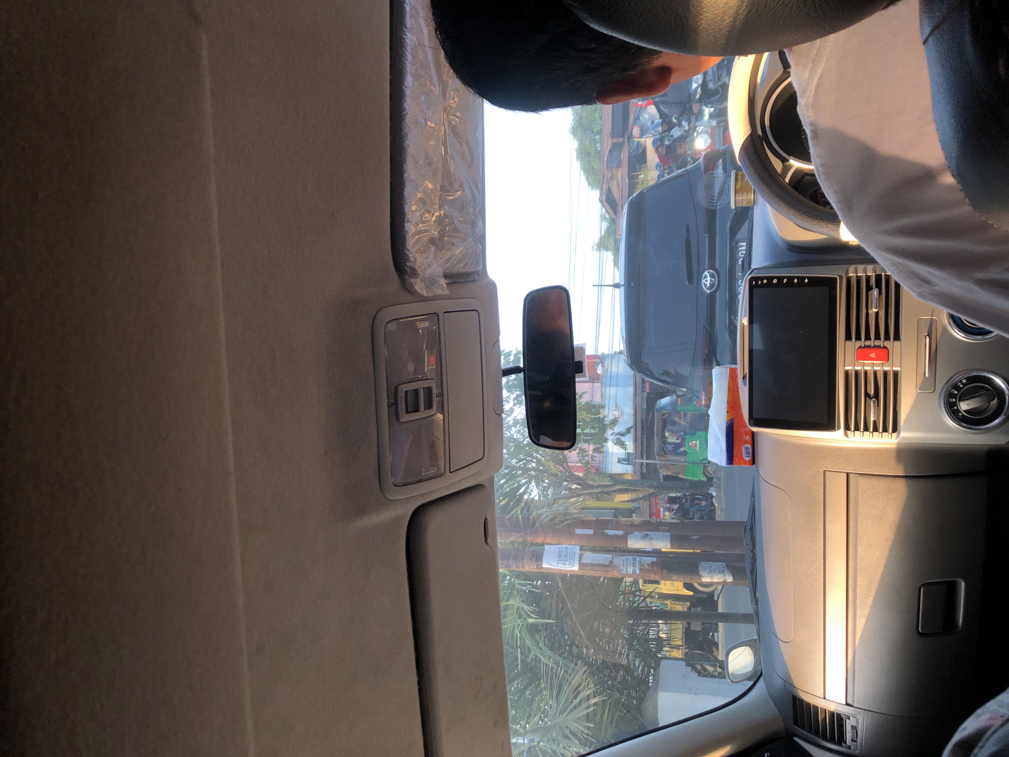 The image is taken from inside a vehicle in the city of Medan, Indonesia, likely on a street with moderate traffic. The perspective is from the passenger seat, looking forward. The dashboard of the car is visible in the foreground, with a touchscreen infotainment system and air vents. A box of tissues is placed on the dashboard. Outside the car, traffic is visible. A black SUV is in front, with motorcycles and a three-wheeled vehicle, possibly an auto-rickshaw, further ahead. Buildings and trees line the street, suggesting an urban or suburban environment. Sunlight is casting strong shadows, indicating it is likely late afternoon or early morning. The sky is bright and slightly overcast. The scene captures a typical moment of being in traffic in a bustling Indonesian city.