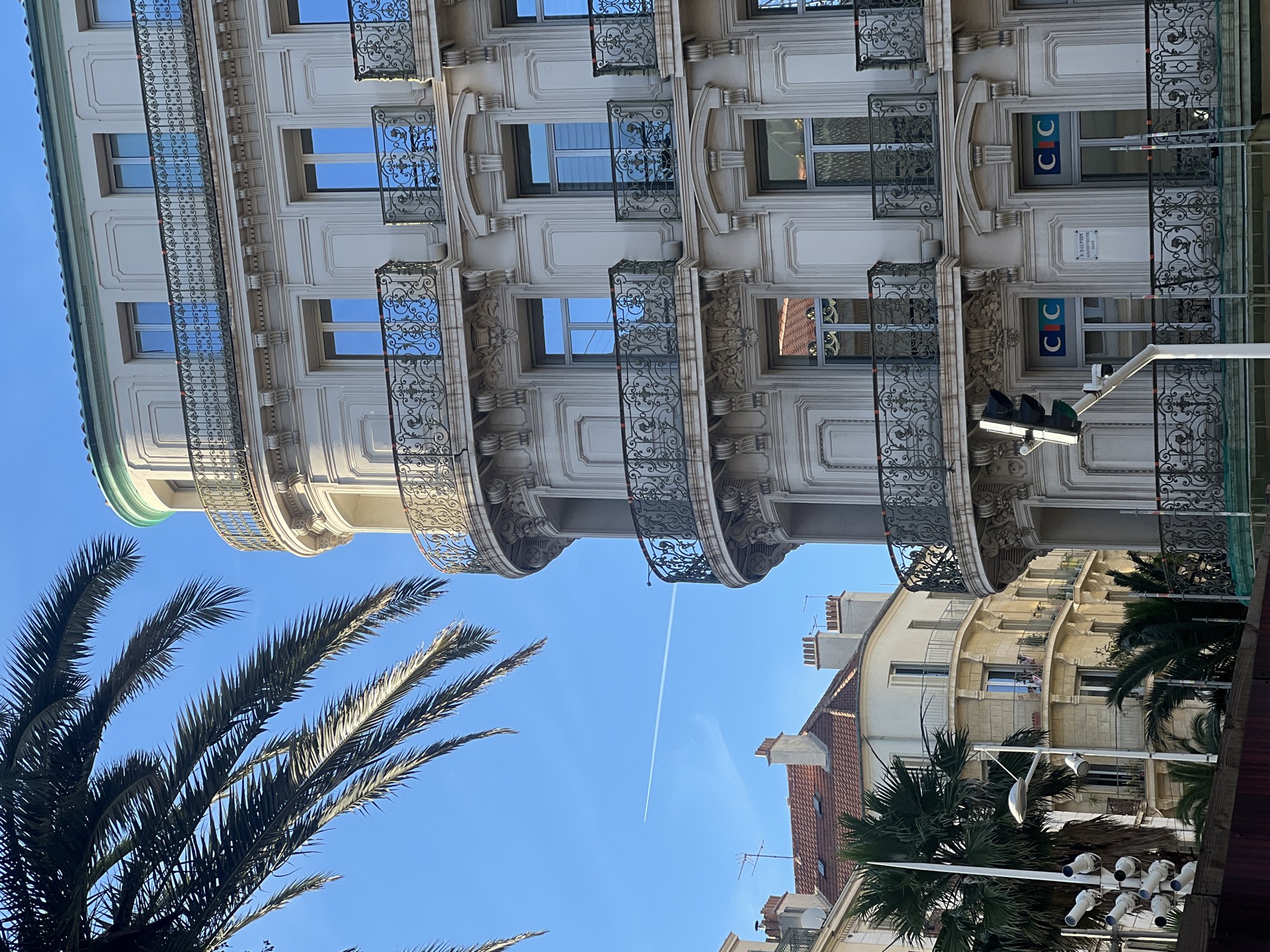 The image depicts a view of a curved building facade with ornate balconies and windows, typical of Haussmannian architecture. The building is light-colored, with decorative iron railings on the balconies. Several windows are visible, reflecting the clear blue sky. In the foreground, a portion of a palm tree frond extends into the frame from the left. In the background, another building with a red tiled roof and chimneys can be seen. A contrail from an airplane is visible in the sky. A traffic light and a surveillance camera are also present in the foreground. The location is identified as Toulon, France.