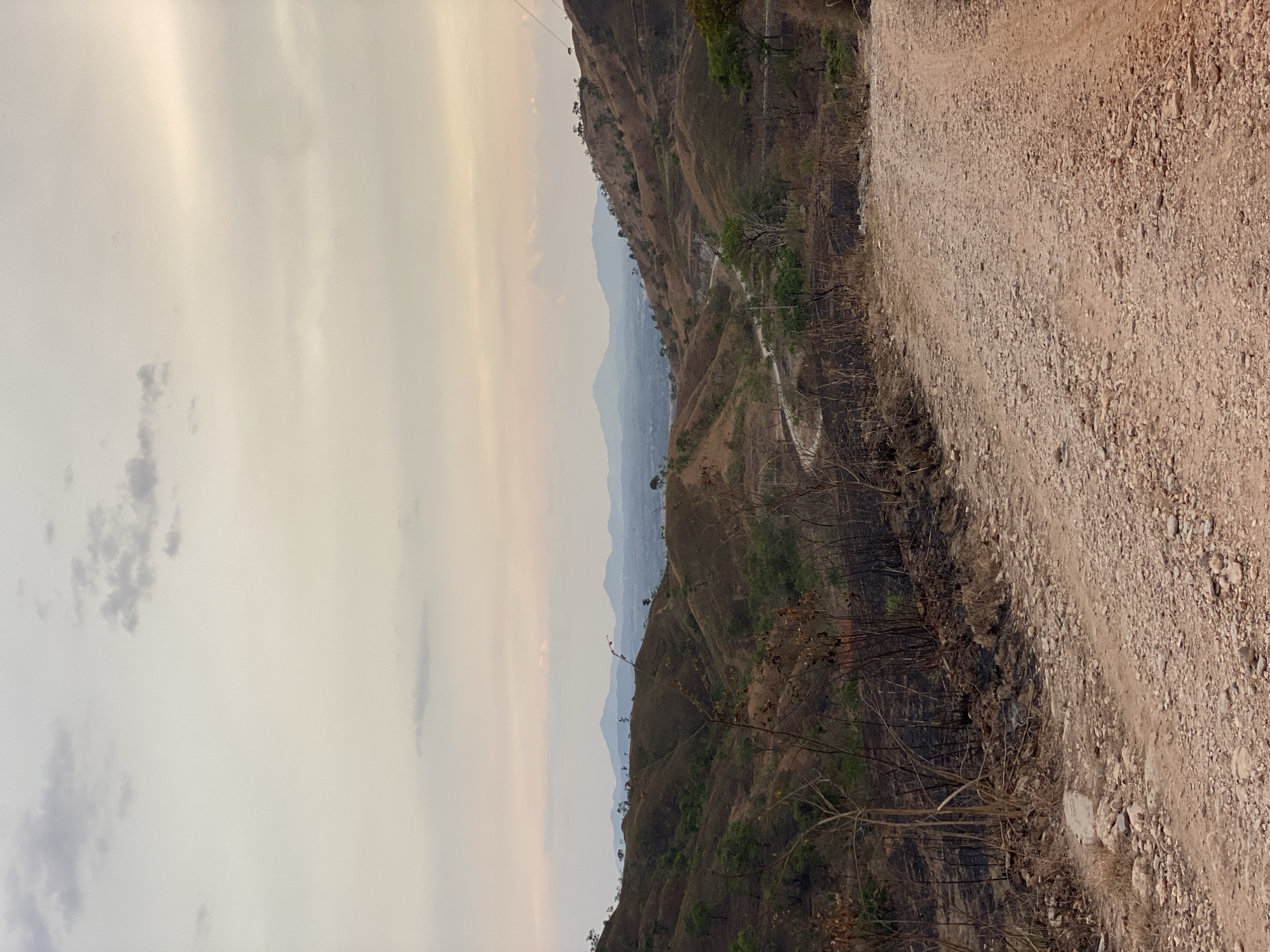 The image shows a landscape in Tinaquillo, Venezuela. A dirt road runs along the right side of the frame. Adjacent to the road, the terrain slopes down into a valley. The vegetation appears to be sparse and burnt, with scattered green patches. Distant hills and a hazy sky form the backdrop. The sky is overcast with light clouds.