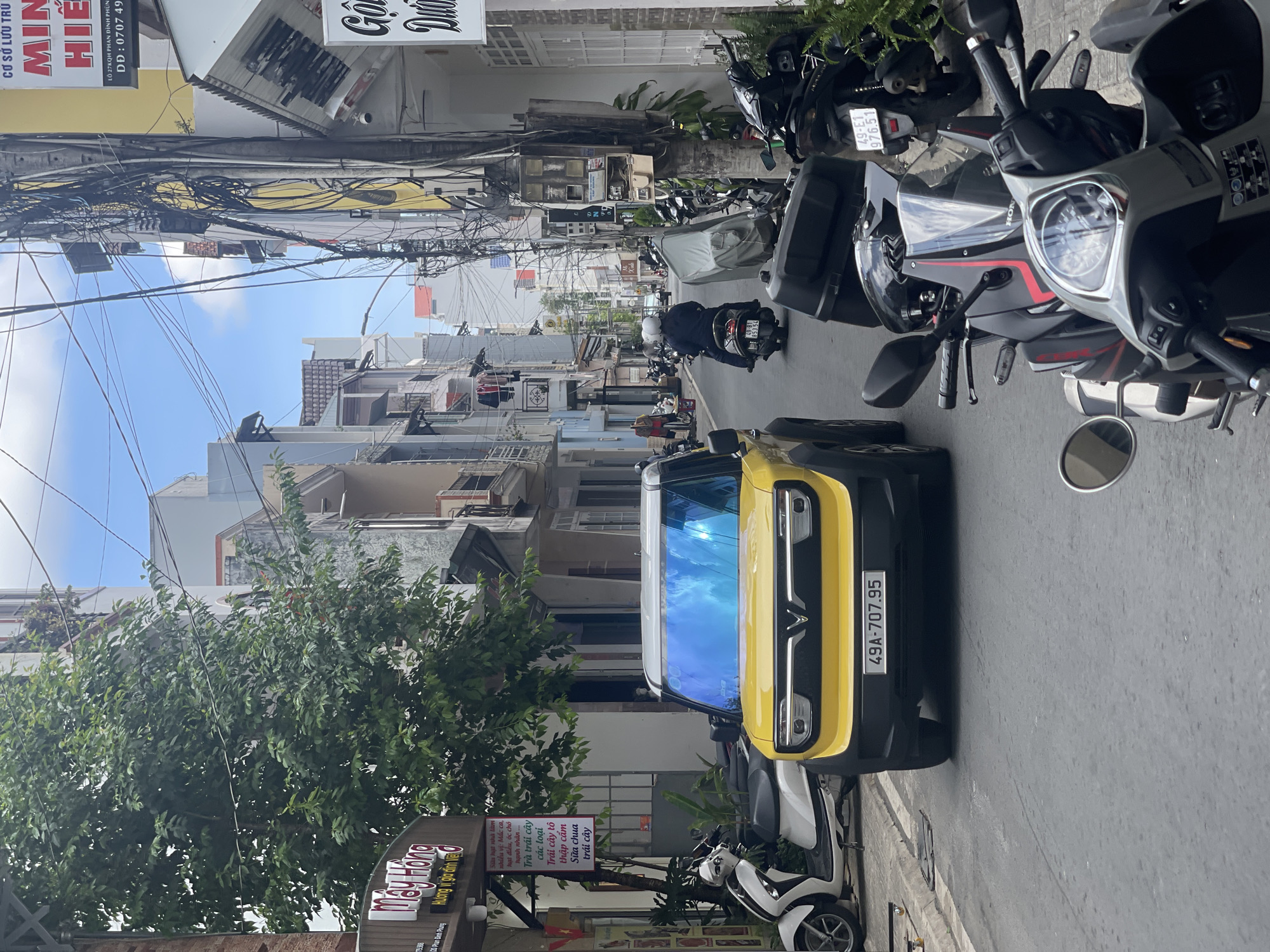The image depicts a street scene in Da Lat, Vietnam. The photograph is taken at street level, with the perspective looking down a narrow road lined with buildings.
A yellow and black VinFast Fadil car is parked on the right side of the street, bearing the license plate "49A-707.95." A white scooter is parked behind it. Numerous parked motorcycles line the right side of the road. A person on a scooter is heading into the distance along the street.
Buildings on both sides of the street are typical of Vietnamese urban architecture, with multiple stories and narrow facades. Utility wires are tangled overhead, obscuring parts of the sky. A tree grows on the left side of the street. Storefront signs are visible, including one for "May Hong" with Vietnamese text, and another for "Minh Hieu" lodging.