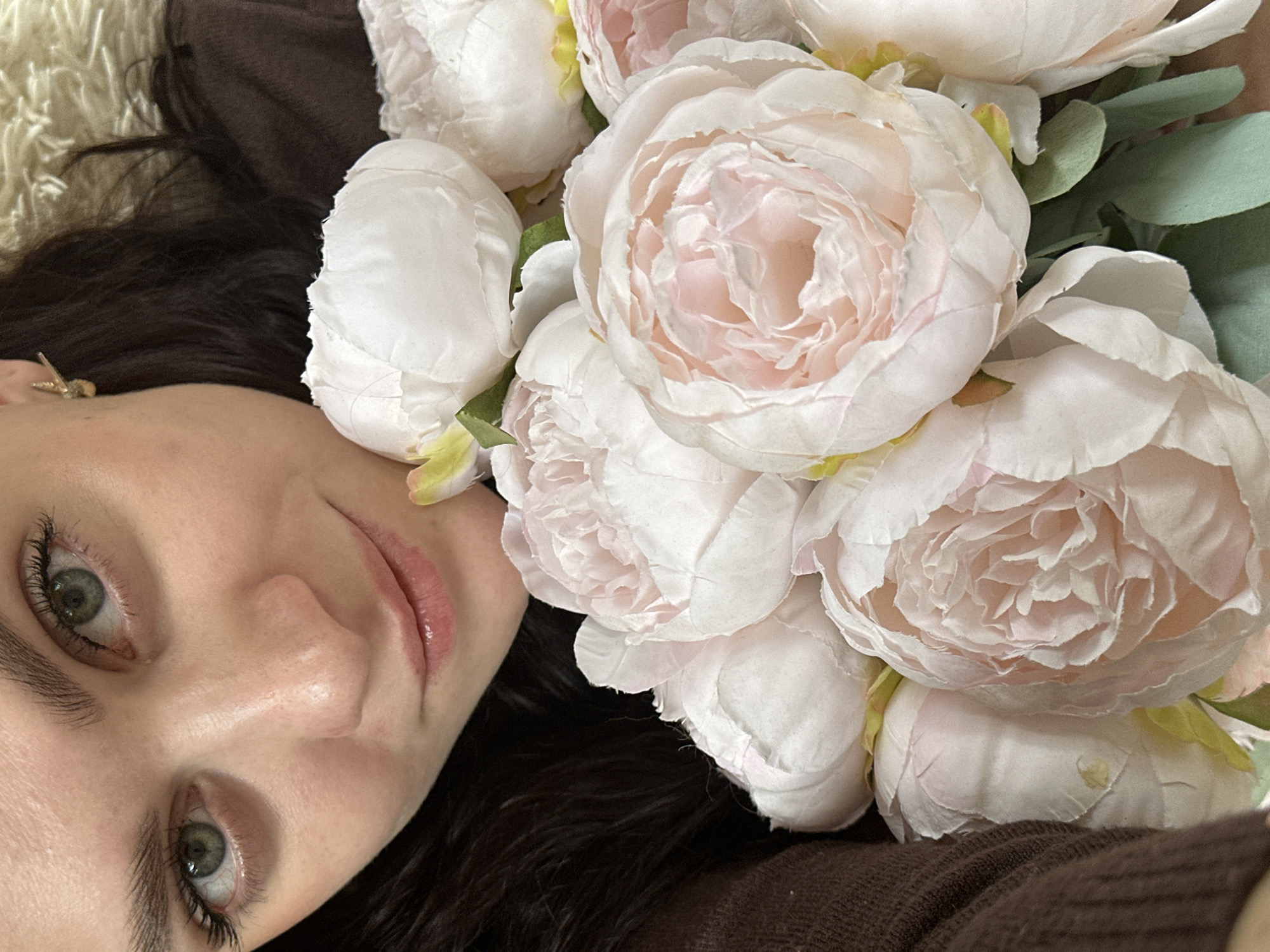 A woman lies on her back with a bouquet of light pink and white peonies obscuring the upper right portion of the frame. She has fair skin, dark brown hair, and green eyes. Her lips appear glossy. A small gold earring is visible in her left ear. She wears a brown garment. The background is slightly out of focus and appears to be a beige, textured surface. It is impossible to determine the location of the image as Chaikovsky, Russia, or any other specific location, based on the provided information.