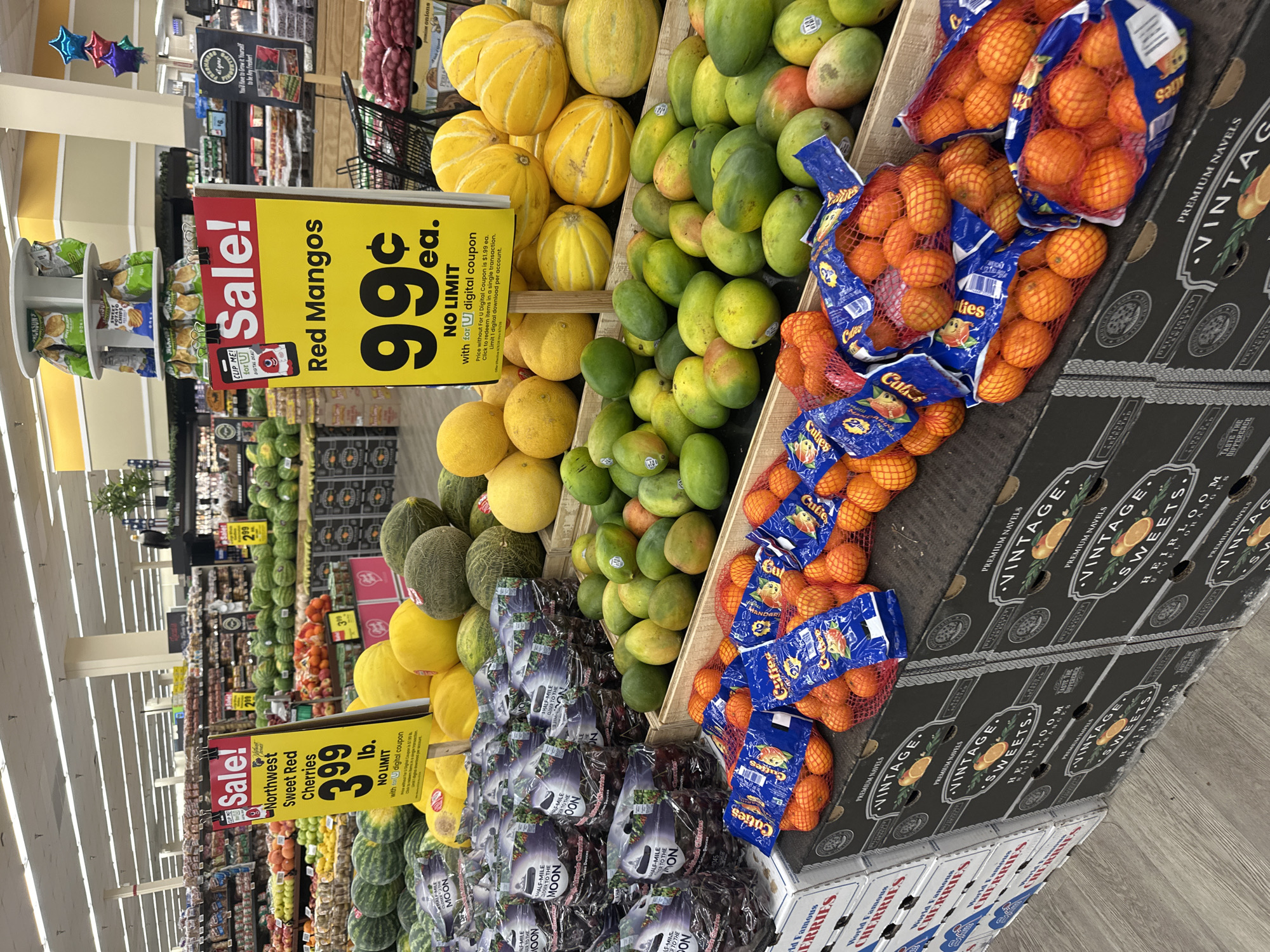 In the produce section of a supermarket in Schaumburg, Illinois, USA, a display of various fruits is prominently featured. In the foreground, several bags of small, orange citrus fruits, labeled "Cuties," are presented in blue netting. Behind these, a large pile of green and yellow mangoes are arranged in wooden crates. To the left, stacks of boxed "World Famous Cherries" are visible. Further into the background, a variety of melons, including watermelons, cantaloupes, and honeydew melons, are on display. Two prominent "Sale!" signs are posted: one for "Northwest Sweet Red Cherries" at $3.99 per pound, and another for "Red Mangos" at 99 cents each with a digital coupon. The environment appears to be a well-lit, indoor retail space with fluorescent lighting and a visible ceiling structure. No individuals are directly captured in the frame.