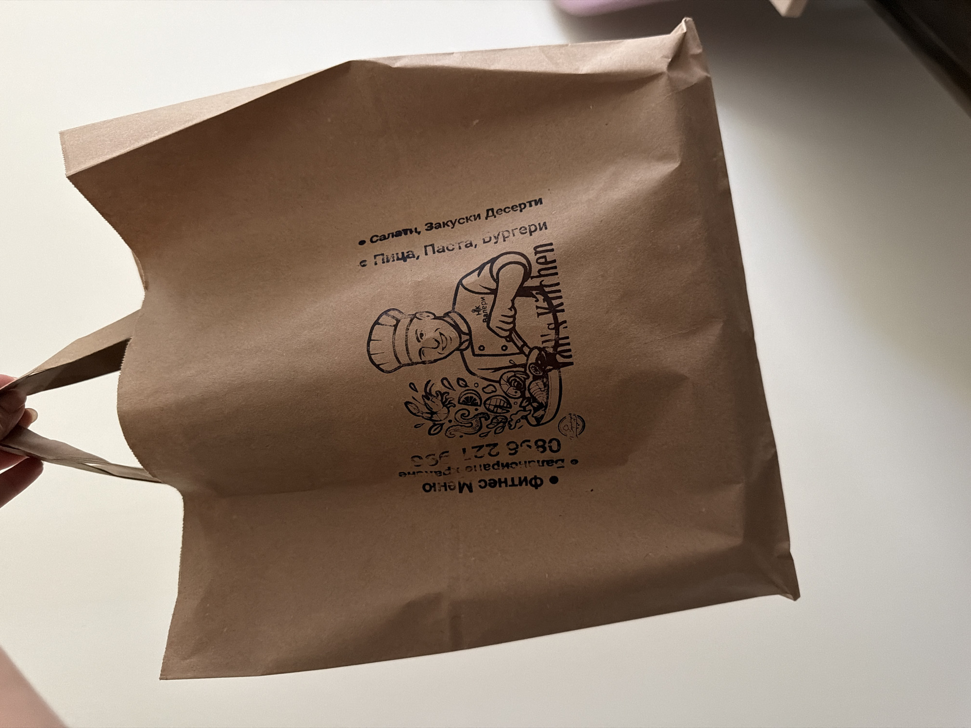 A brown paper shopping bag is held by a person's hand, out of frame at the top. The bag has a logo and text printed on it. The logo depicts a chef with a smiling face, wearing a chef's hat and apron, holding a spatula and pointing to a pile of food. The text on the bag is in Bulgarian and includes "Фитнес Меню" (Fitness Menu), "Балкангранд.Радославе" (Balkangrand.Radoslave), and a phone number "0896 221 555". Another line of text reads "с Пица, Паста, Бургери" (with Pizza, Pasta, Burgers) and "Салати, Закуски Десерти" (Salads, Appetizers, Desserts). The text "Yan's Kitchen" is also visible below the chef illustration. The bag is against a plain, light-colored background, possibly a wall. The location is Pazardzhik, Bulgaria.