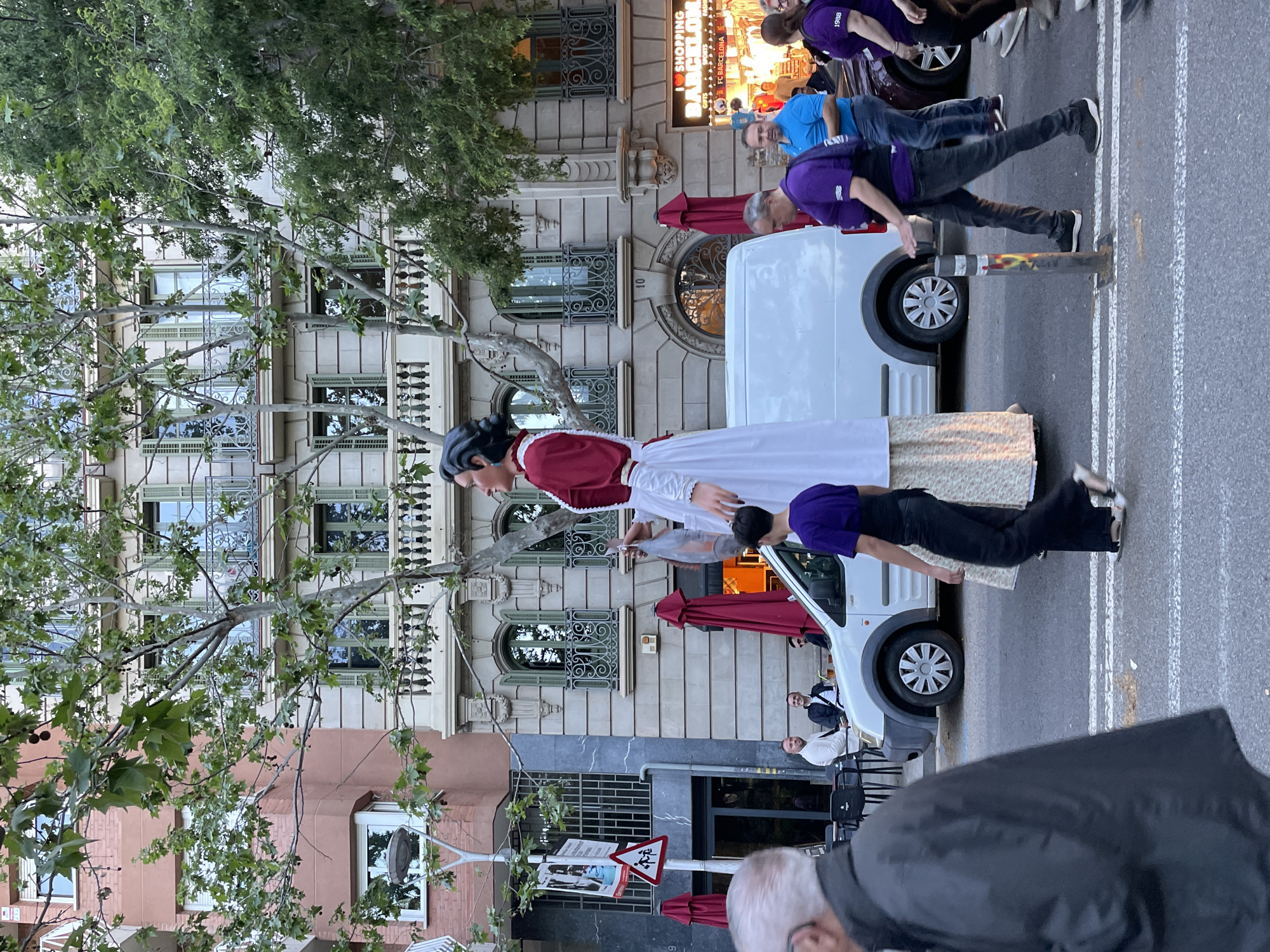 The image shows a street scene in Barcelona, Spain. The focus is on a large traditional Catalan figure (likely a "gegant") being carried by a person wearing a purple shirt. A group of people, also in purple shirts, accompany the figure. In the background is a white van and a building with ornate windows. Street signs and a cafe are also visible. The overall impression is a cultural event or celebration taking place on a city street.