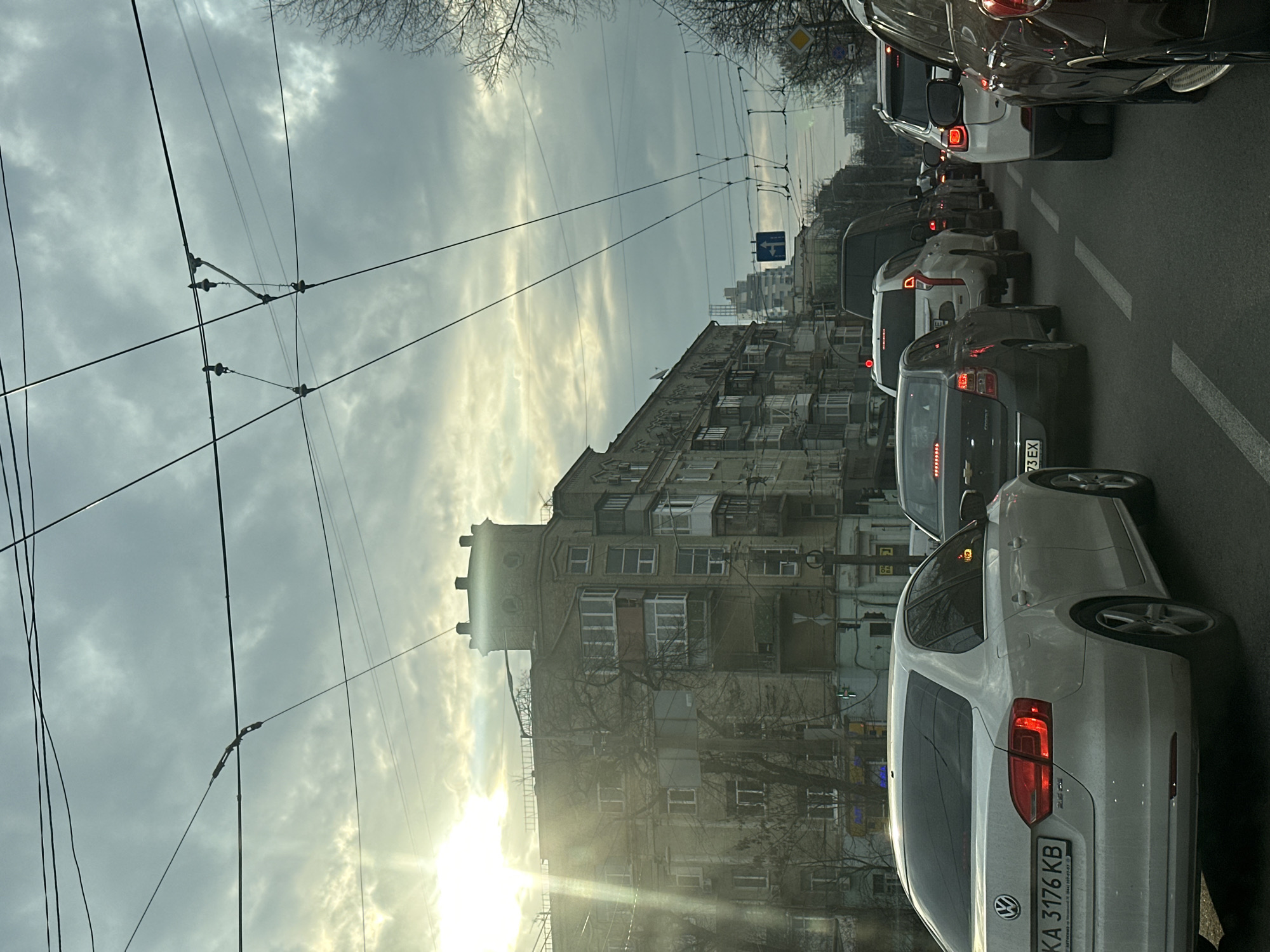 The image shows a traffic jam on a street in Kyiv, Ukraine. The street is lined with buildings, including an older residential building with a distinctive tower. Overhead are trolleybus wires. The sky is overcast with the sun low on the horizon. The traffic consists of several cars, including a white Volkswagen with a license plate "KA 3176 KB," a dark-colored Chevrolet, and other various vehicles.