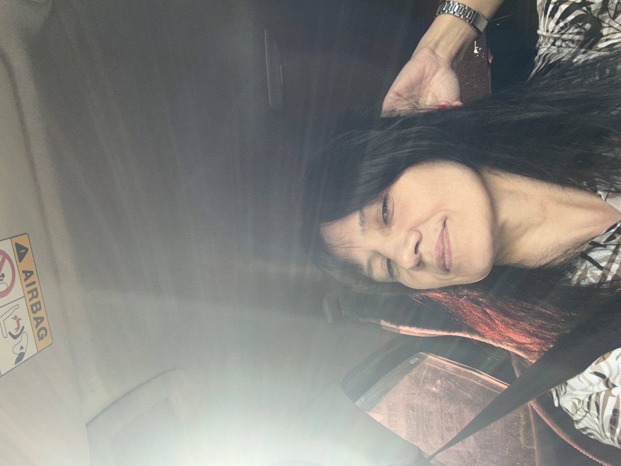 The image shows a woman in a car. She is wearing a patterned shirt and a silver watch. She is smiling slightly. The car interior is visible, including an airbag warning label and a seatbelt. The location is unknown.