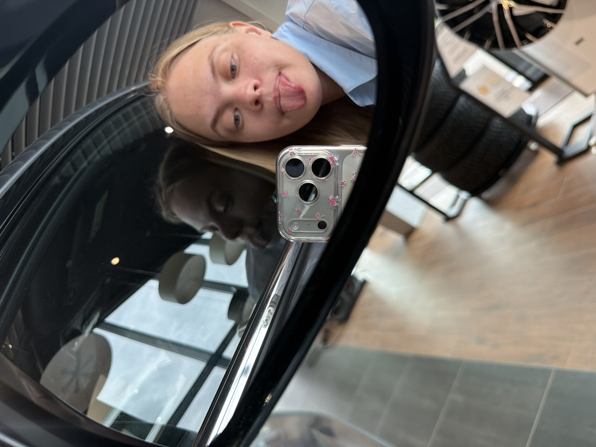 A person is visible in a reflective surface, seemingly a car window or mirror. The person is fair-skinned with blonde hair and wearing a light blue collared shirt. Their tongue is sticking out, suggesting a playful or silly pose. A smartphone with a clear case adorned with small floral designs is held up to take the picture, obscuring part of the face. The reflection in the surface includes elements of a modern interior, likely a showroom or dealership. Objects visible include furniture (possibly chairs with patterned upholstery), lighting fixtures, and large windows with multiple panes. The environment appears well-lit and clean. There is no information available in the image to suggest the location is Izhevsk, Russia.