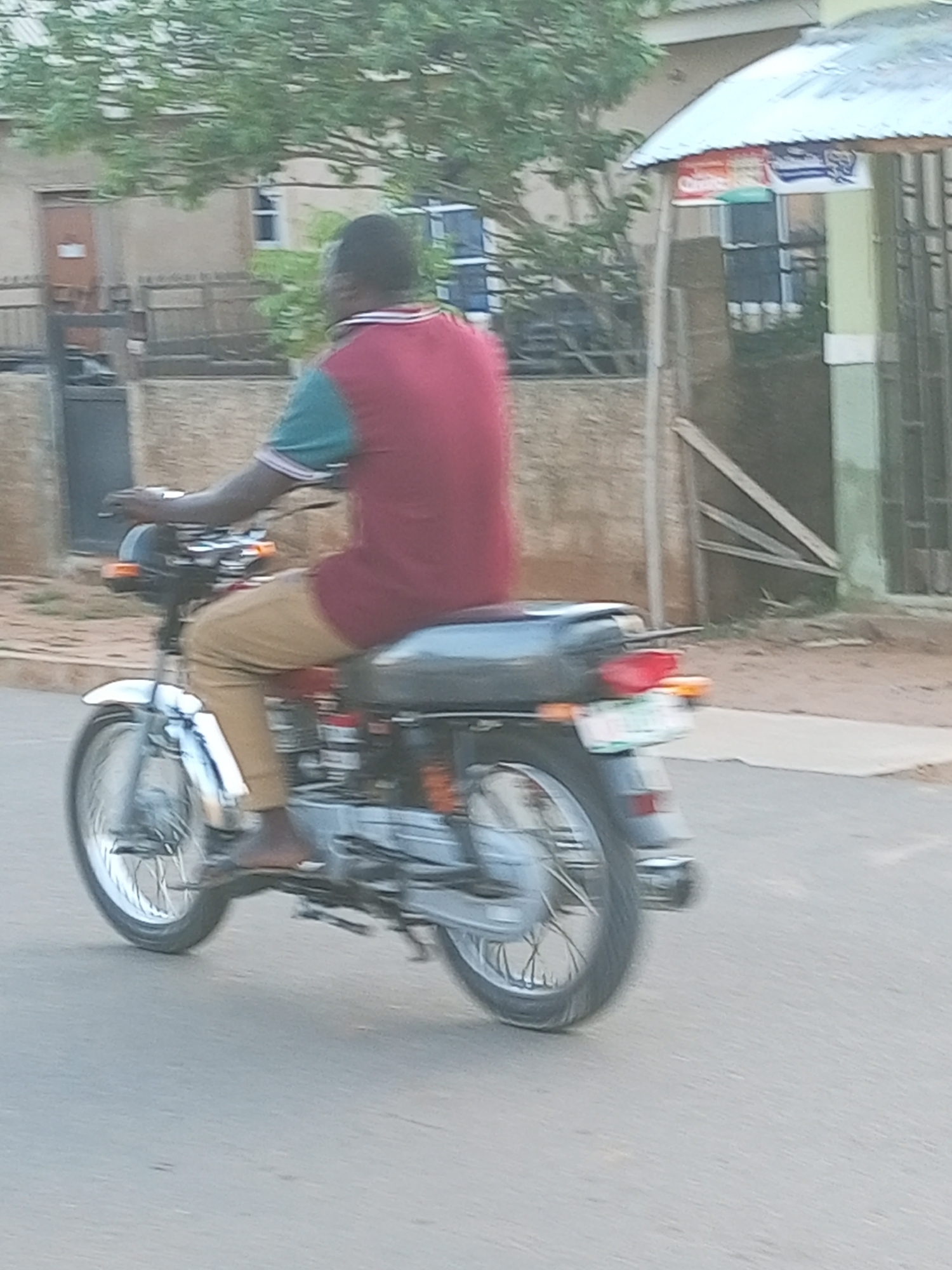 In Tempure, Nigeria, a man with dark skin is riding a silver and black motorcycle. He is wearing a red and teal shirt with a white collar and tan pants. The man is barefoot. The motorcycle has a license plate that is partially visible, displaying green and white text. The motorcycle has a red taillight and orange turn signals. The background shows a building with a light green facade and a white roof, possibly a shop. A tree with green leaves is visible in the upper left corner of the image. The ground is paved with asphalt. The image is slightly blurred, suggesting movement.