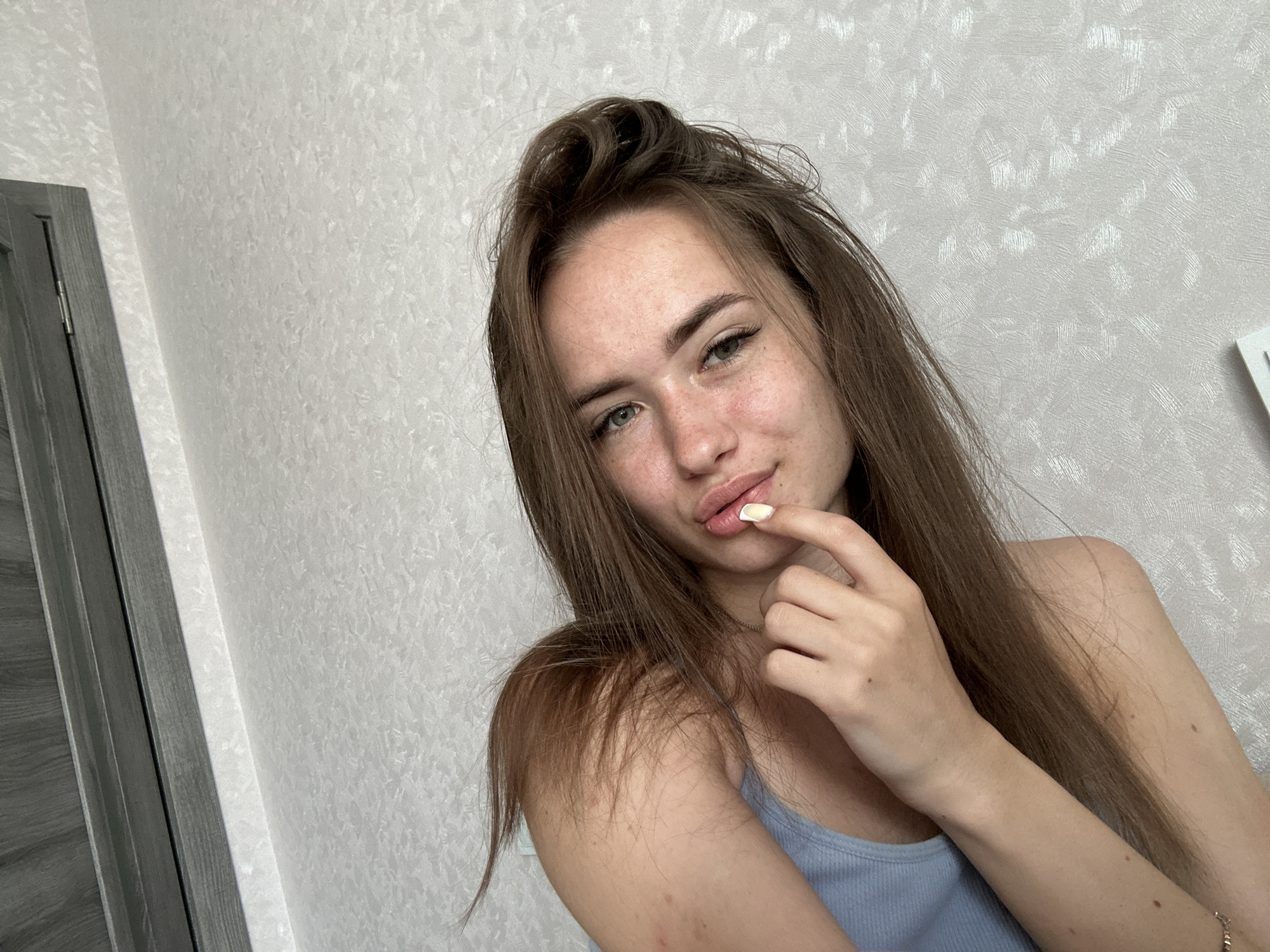 A young woman with long, brown hair and light-colored eyes is captured in a selfie. She is looking directly at the camera, with her right hand touching her lips. She is wearing a light blue tank top and her skin has a scattering of freckles. The background consists of a textured, light-colored wall and a portion of a grey door on the left side of the frame.