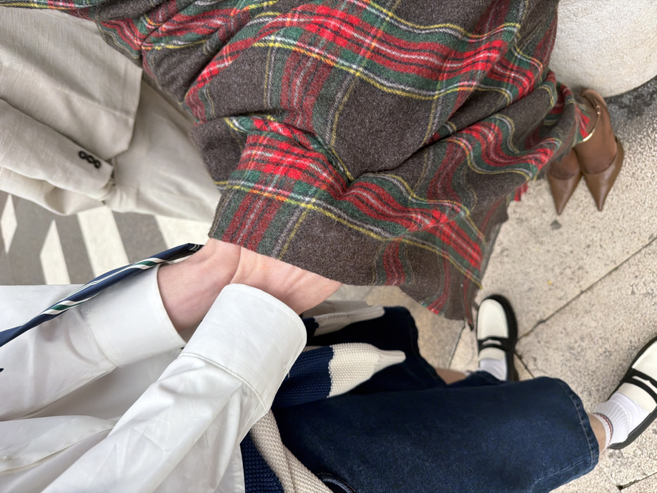 The image captures a close-up of three individuals on a paved urban street in Milan, Italy, with a pedestrian crossing visible in the background. In the immediate foreground, two individuals are partially depicted. The person on the left wears a white long-sleeved shirt with folded cuffs, dark blue denim trousers or a wide-leg skirt, white socks with a thin red stripe at the top, and black and white flat shoes. A blue and white striped knit garment is draped over their left forearm. The person on the right is attired in a dark brown plaid garment featuring prominent red, green, and thin yellow lines, and brown pointed-toe heels with a gold ankle strap. Their left hand is positioned over or directly adjacent to the left individual's right hand. In the blurred mid-ground, a third individual is partially visible, wearing a light beige suit jacket with three dark buttons on the cuff and matching trousers. The ground beneath the subjects is a light-colored paved surface with visible seams. In the out-of-focus background, white stripes of a pedestrian crosswalk are discernible on a darker road surface.