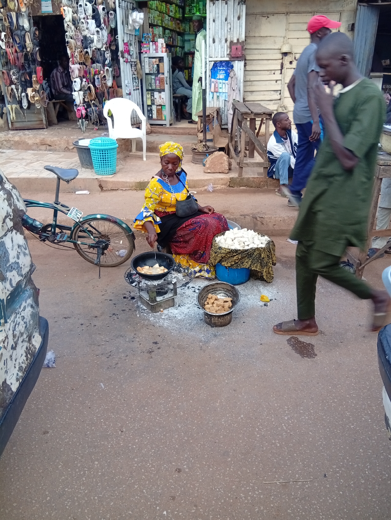 In Bukuru, Nigeria, a street scene unfolds with a woman in a vibrant yellow and blue patterned dress, her head covered by a yellow headscarf, frying food in a black pan placed on a charcoal stove. She is positioned on the dusty, paved ground, with a bicycle to her left and a blue bin with a light blue lid beside her. A pile of what appears to be white diced food is visible behind her, covered by a patterned cloth. To her right, a metal sieve contains pieces of fried food. A man in a green robe walks past in the foreground. In the background, a shop displays numerous shoes and other items. Several men are also visible, some seated and others standing. The environment suggests a typical market or street-side vending area.