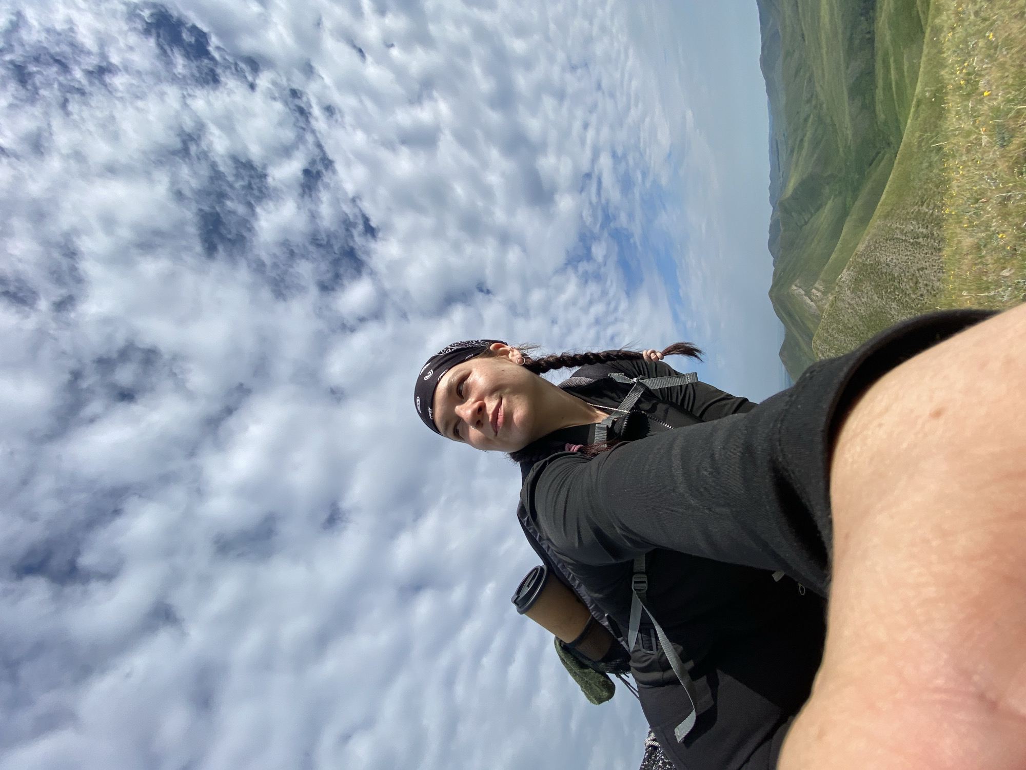 A young woman with braided hair and a bandana is taking a selfie while hiking in a mountainous region. She is wearing a black jacket and a backpack. The landscape behind her features rolling green hills under a cloudy sky. The location is Verkhnyaya Chernorechka, Russia.