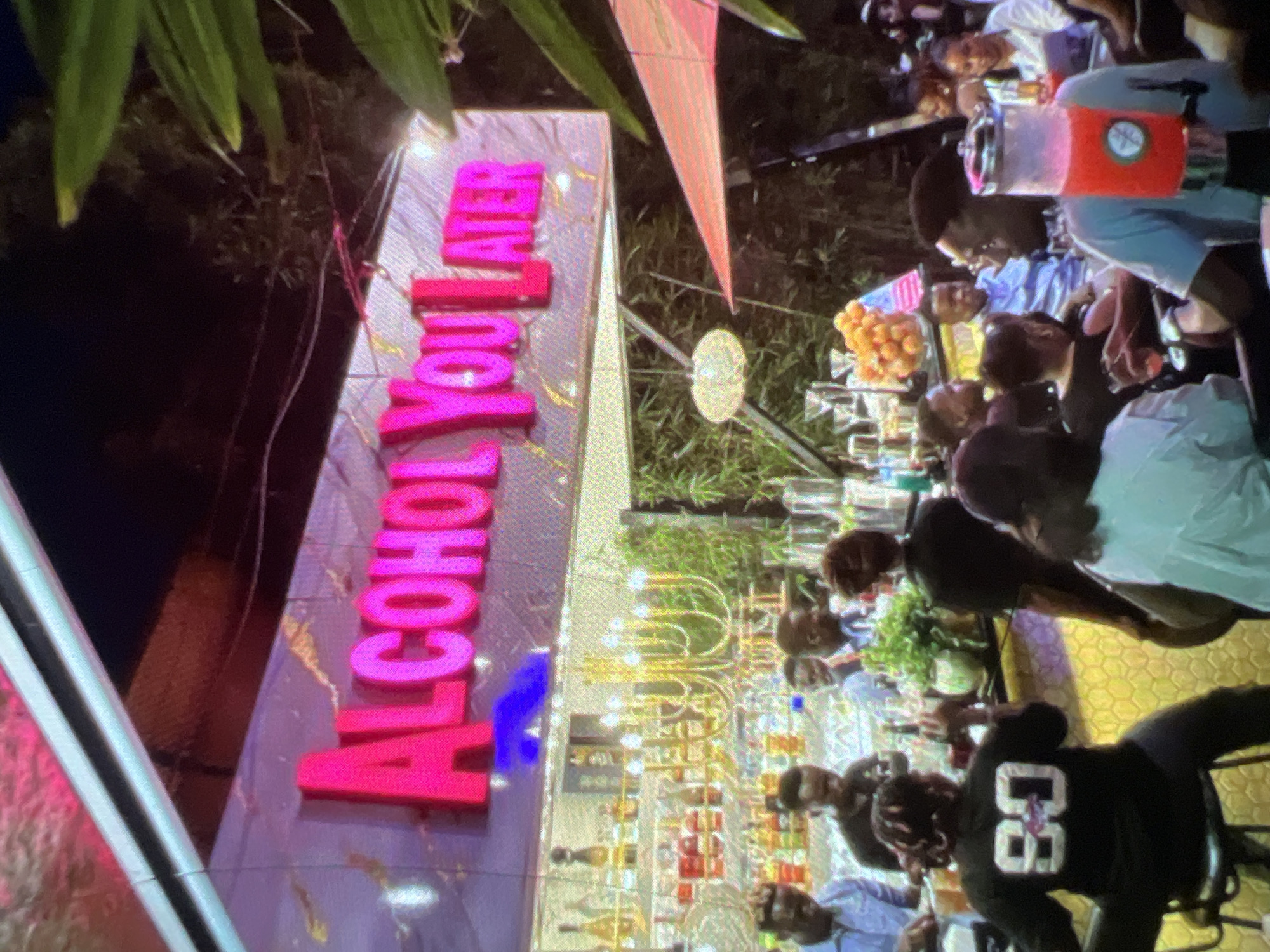 The image depicts a nighttime scene at an outdoor bar or lounge in Lagos, Nigeria, identified by the "ALCOHOL YOU LATER" neon sign. The foreground features a row of people seated or standing at a bar counter. On the far left, a person wearing a black t-shirt with the number "80" on the back is visible. Behind the bar, shelves are stocked with various bottles of alcohol and glassware. A prominent gold chandelier hangs above the bar area. Further back, several individuals are seated at tables, with some holding drinks. An orange-colored drink dispenser is visible in the middle ground to the right. The environment appears to be a well-lit, possibly tropical, setting with foliage and umbrellas indicating an outdoor space.