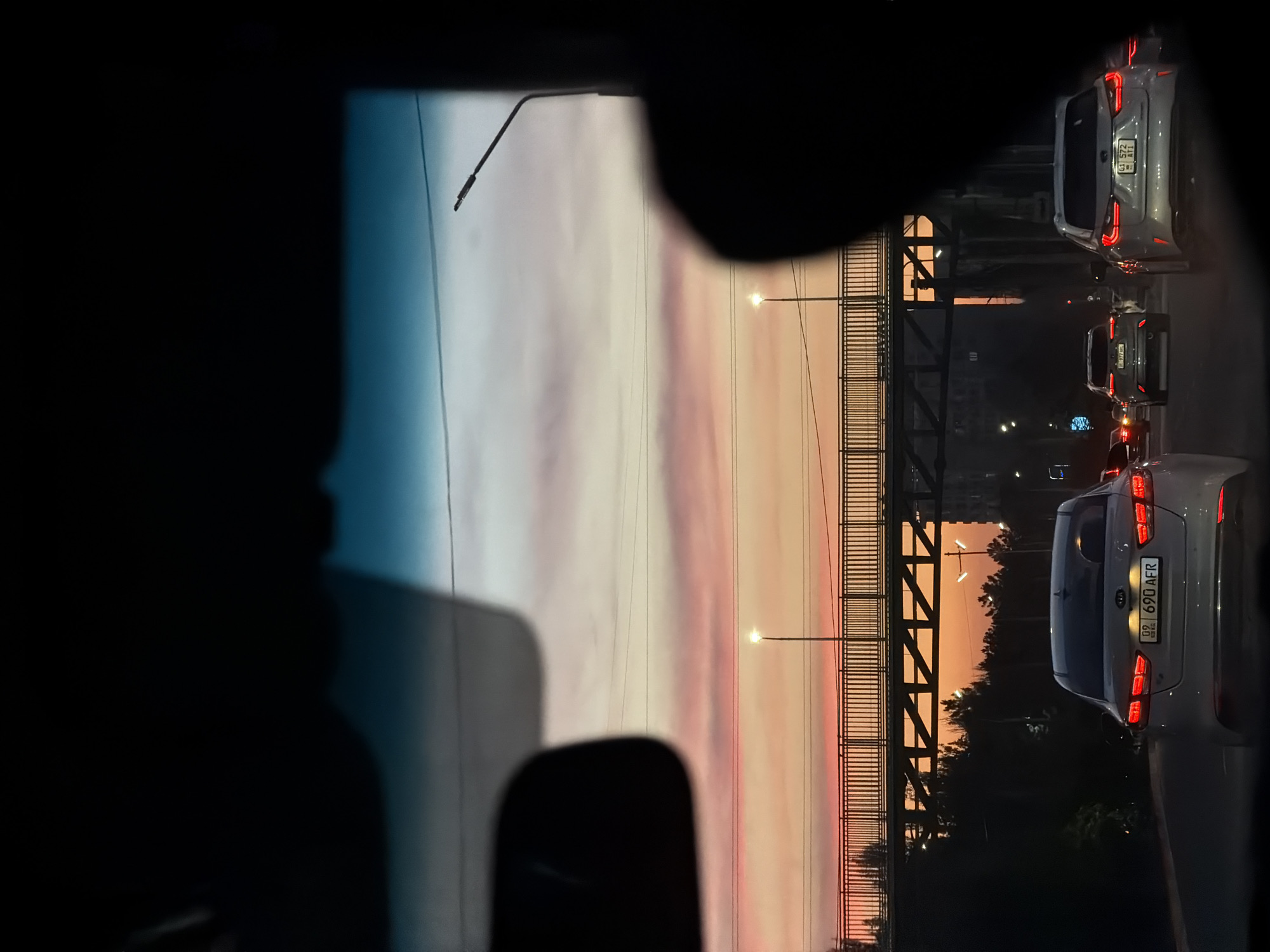 Captured from within a vehicle, the image presents a view of a street in Bishkek City, Kyrgyzstan. The scene features a multi-lane road, a pedestrian overpass, and a backdrop of buildings. Several cars are positioned in front of the camera vehicle, their rear lights visible. The sky displays a gradient of colors, indicating either sunrise or sunset. Streetlights illuminate the scene, adding to the ambient lighting. The photograph is taken at ground level, and the focus is directed towards the vehicles and the overpass in the distance.