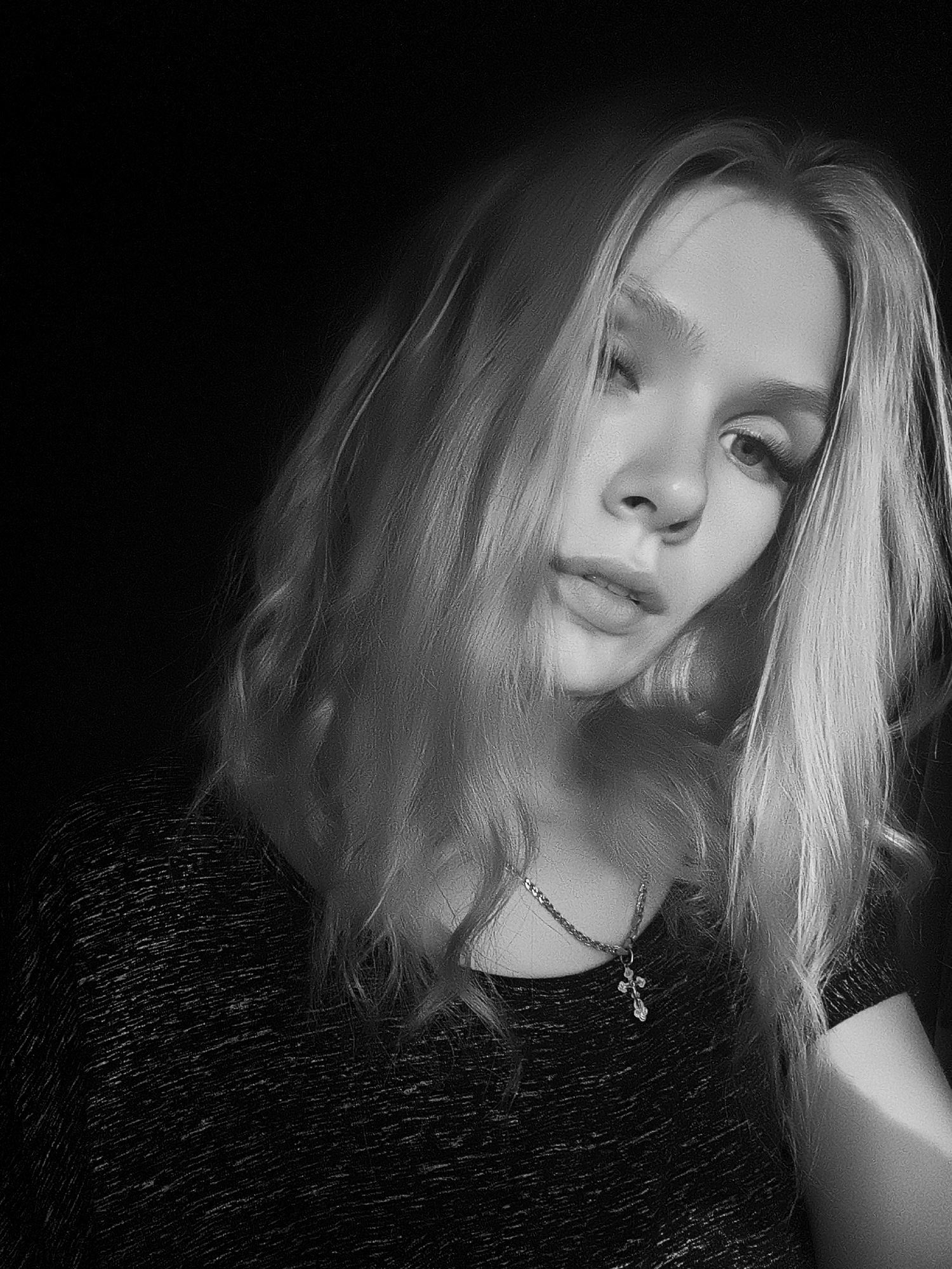 A black and white medium close-up shot shows a young woman with long, blonde, wavy hair against a dark, plain background. The lighting creates strong contrasts, with the right side of her face and hair illuminated by a bright light source, while the left side is cast in shadow. She is looking towards the camera with her head tilted. Her eyes are open, with one slightly more visible than the other. Her lips are slightly parted, and her expression is contemplative or perhaps a little melancholic. She is wearing a dark, textured shirt and a delicate silver chain necklace with a small cross pendant. The overall mood is intimate and artistic, emphasizing the subject's features and the dramatic play of light and shadow. The setting appears to be indoors, with no discernible background details other than darkness. The time of day and weather are not indicated. There is no visible text in the image.