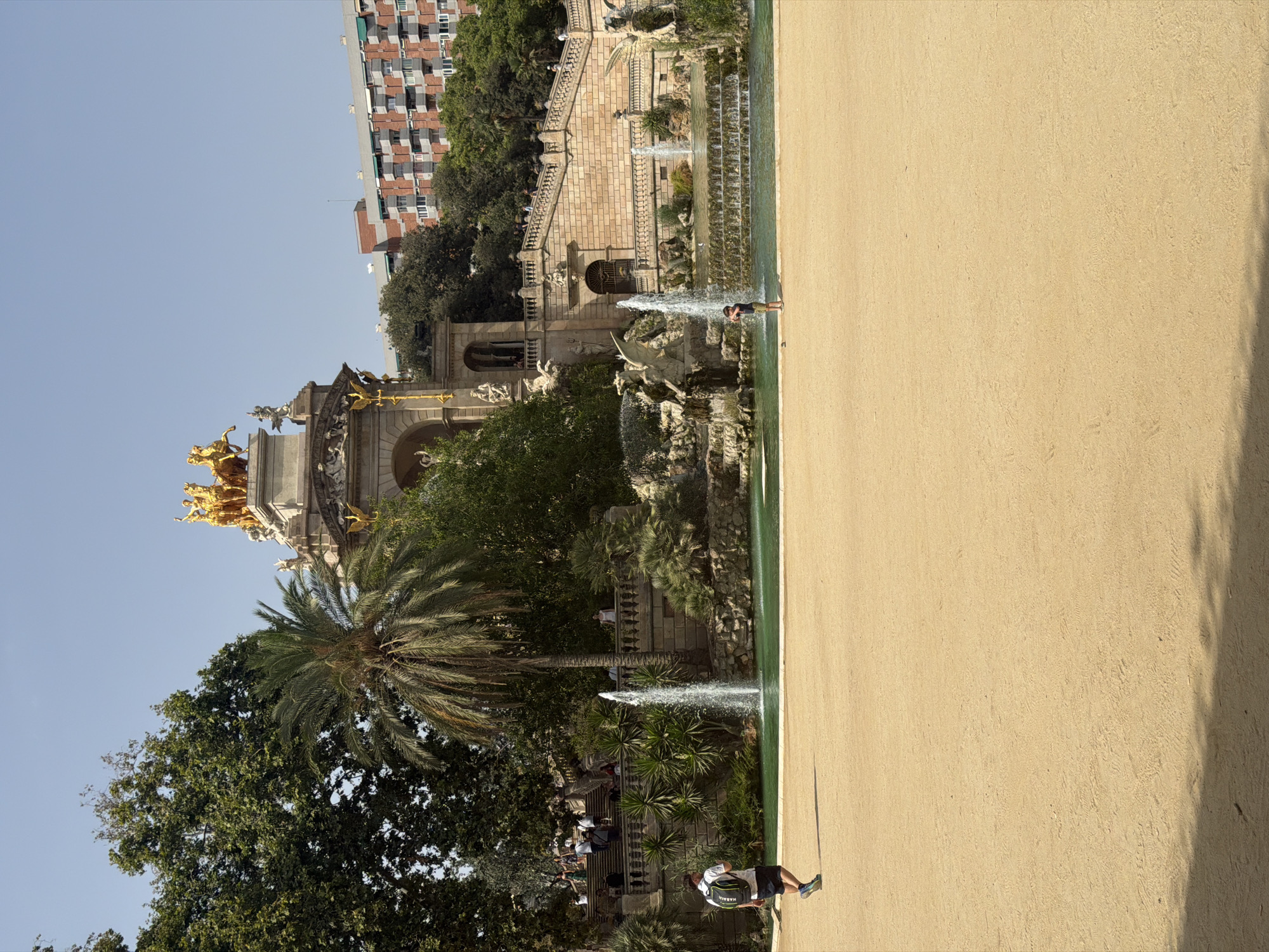 The image depicts a sunny day at Parc de la Ciutadella in Barcelona, Spain. In the foreground, a person is walking across a large sandy area, casting a shadow. Behind them, a pond with fountains and lush greenery is visible. Further back, a grand monument with a golden chariot on top stands out against the blue sky. Several people are seen walking up a wide staircase leading to the monument. In the background, apartment buildings and trees frame the scene. The overall atmosphere is one of a pleasant day in a public park, with both natural and architectural elements contributing to the picturesque setting.